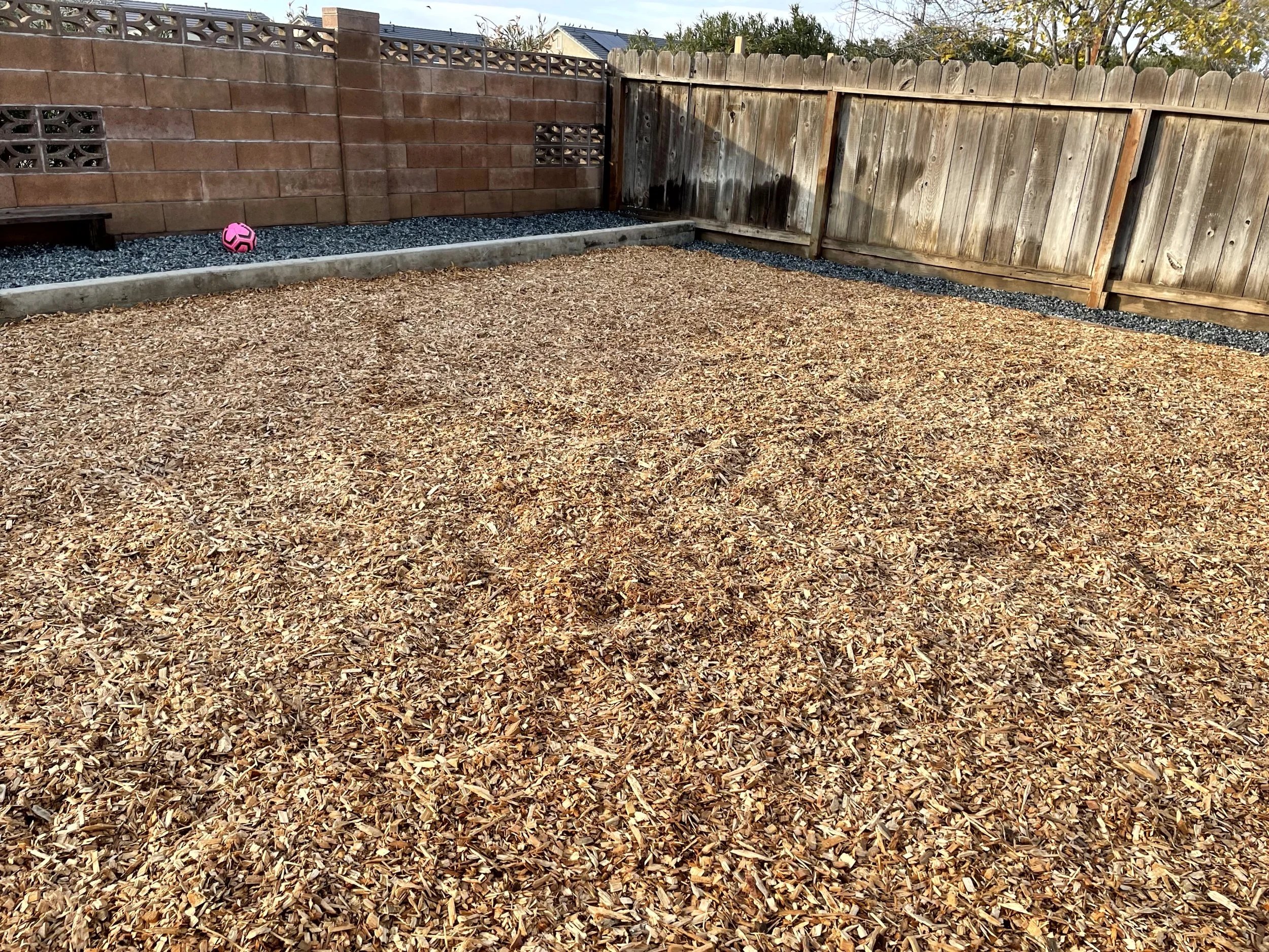 Backyard with wood mulch ground, wooden and brick fencing, and a pink soccer ball near the left fence.