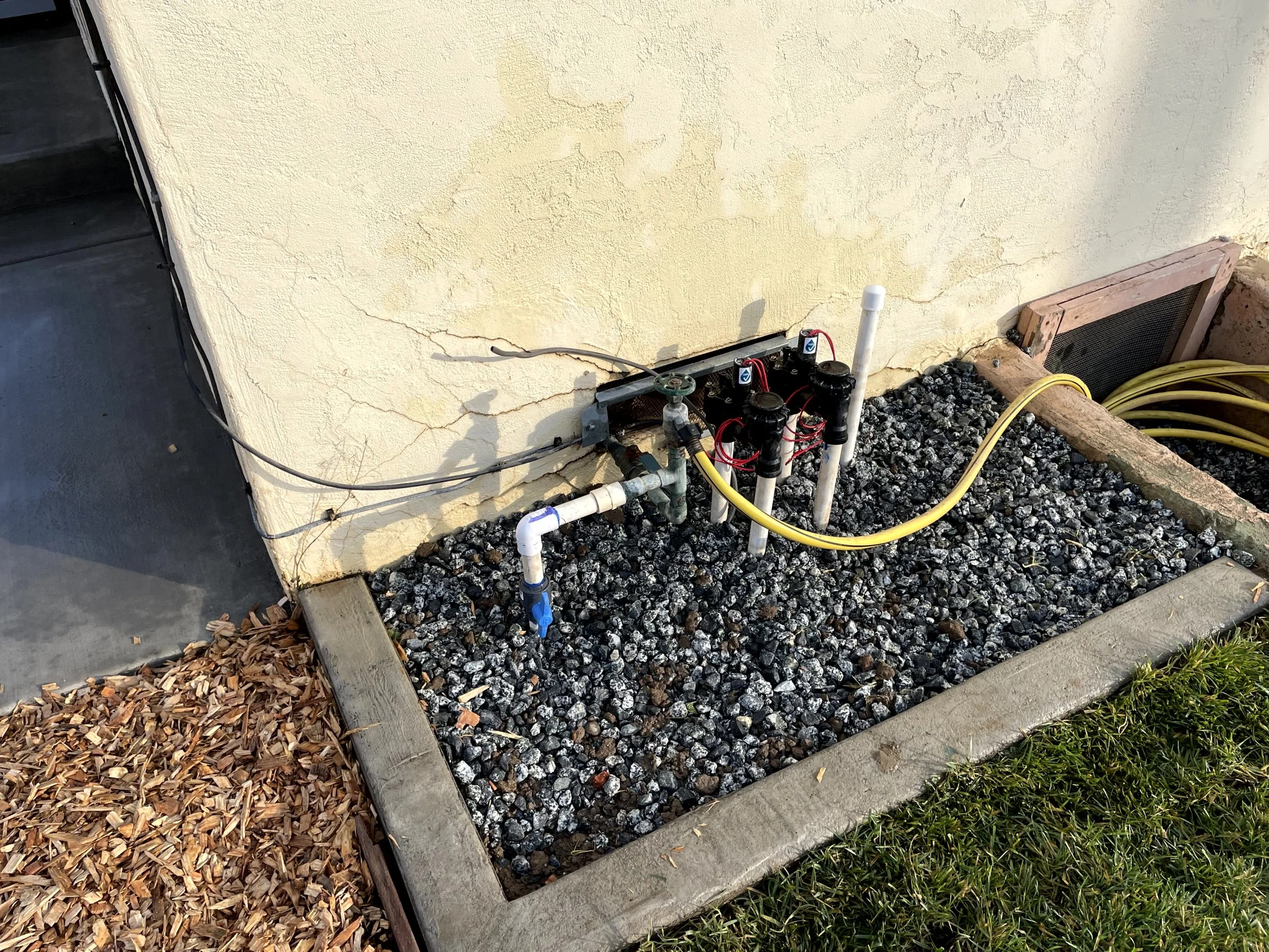 Irrigation control system with valves, pipes, and electrical wiring installed outside a building, surrounded by gravel and framed with wood and concrete border.
