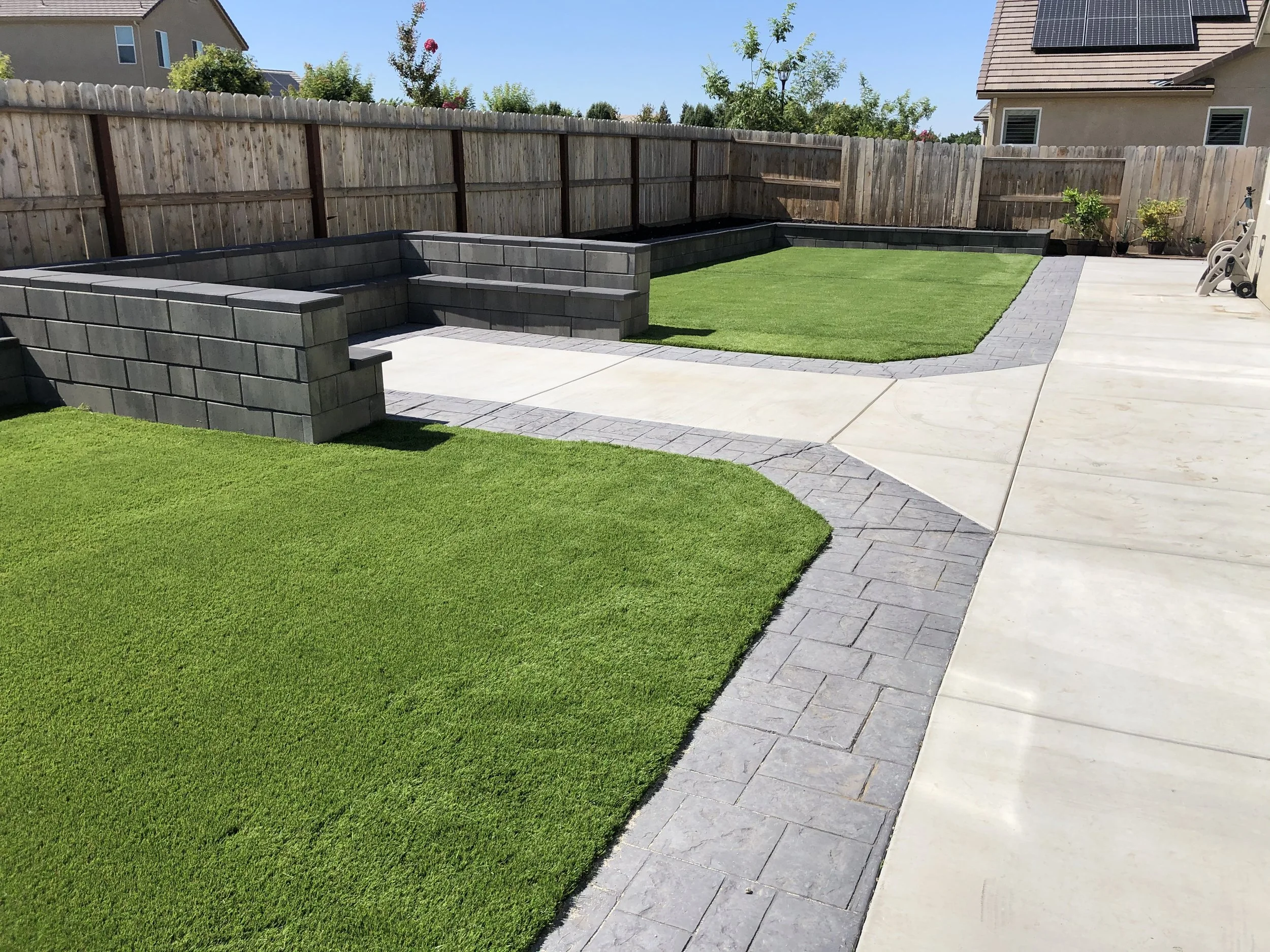A backyard with a fenced perimeter, a small lawn, paved pathways, and a section of patio with a lawn chair.