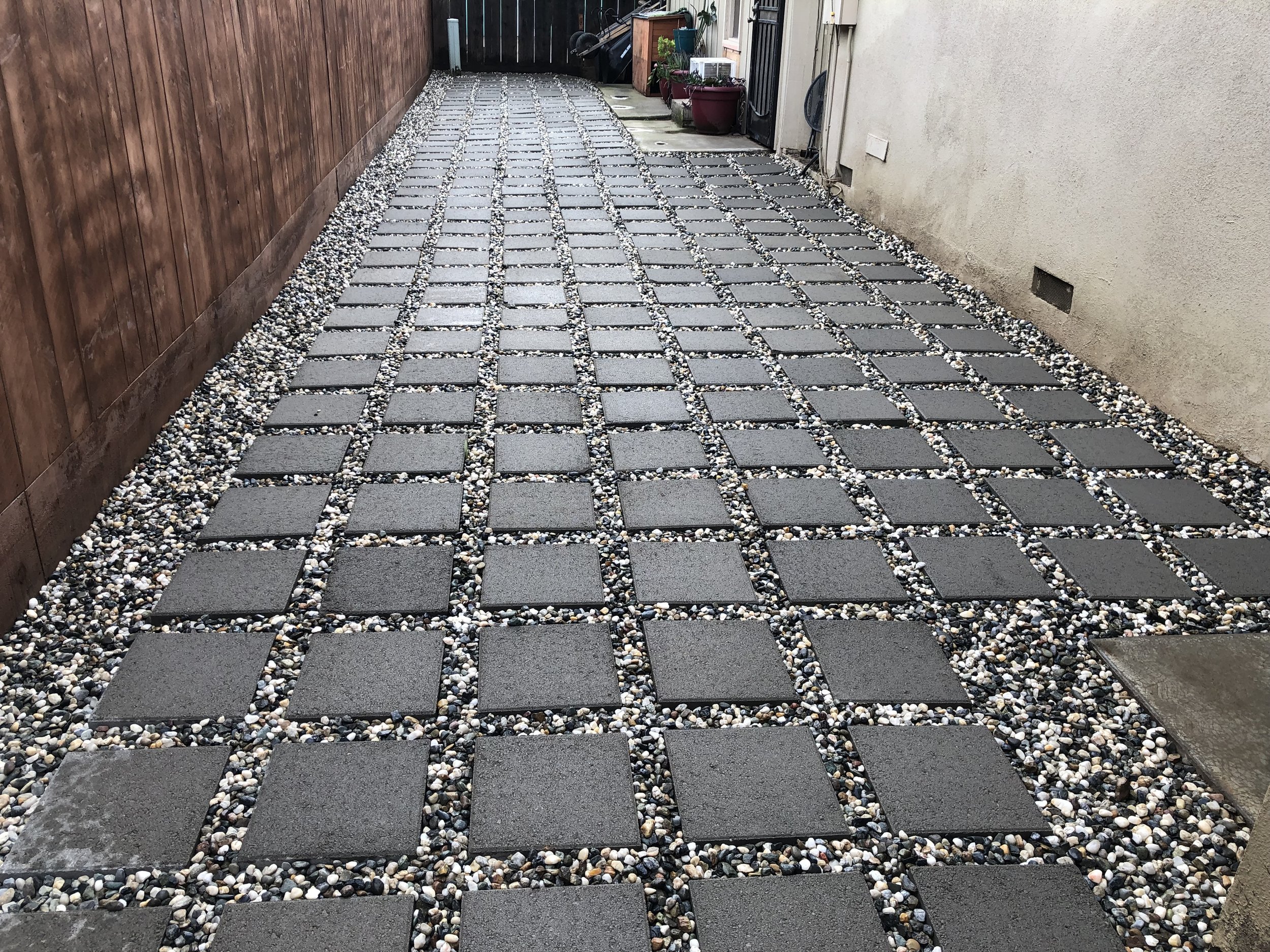 A narrow backyard or side yard pathway paved with square gray concrete tiles, bordered by small multicolored pebbles on both sides, with a wooden fence on the left and a beige wall with a small ventilation opening on the right, leading to a gate in t
