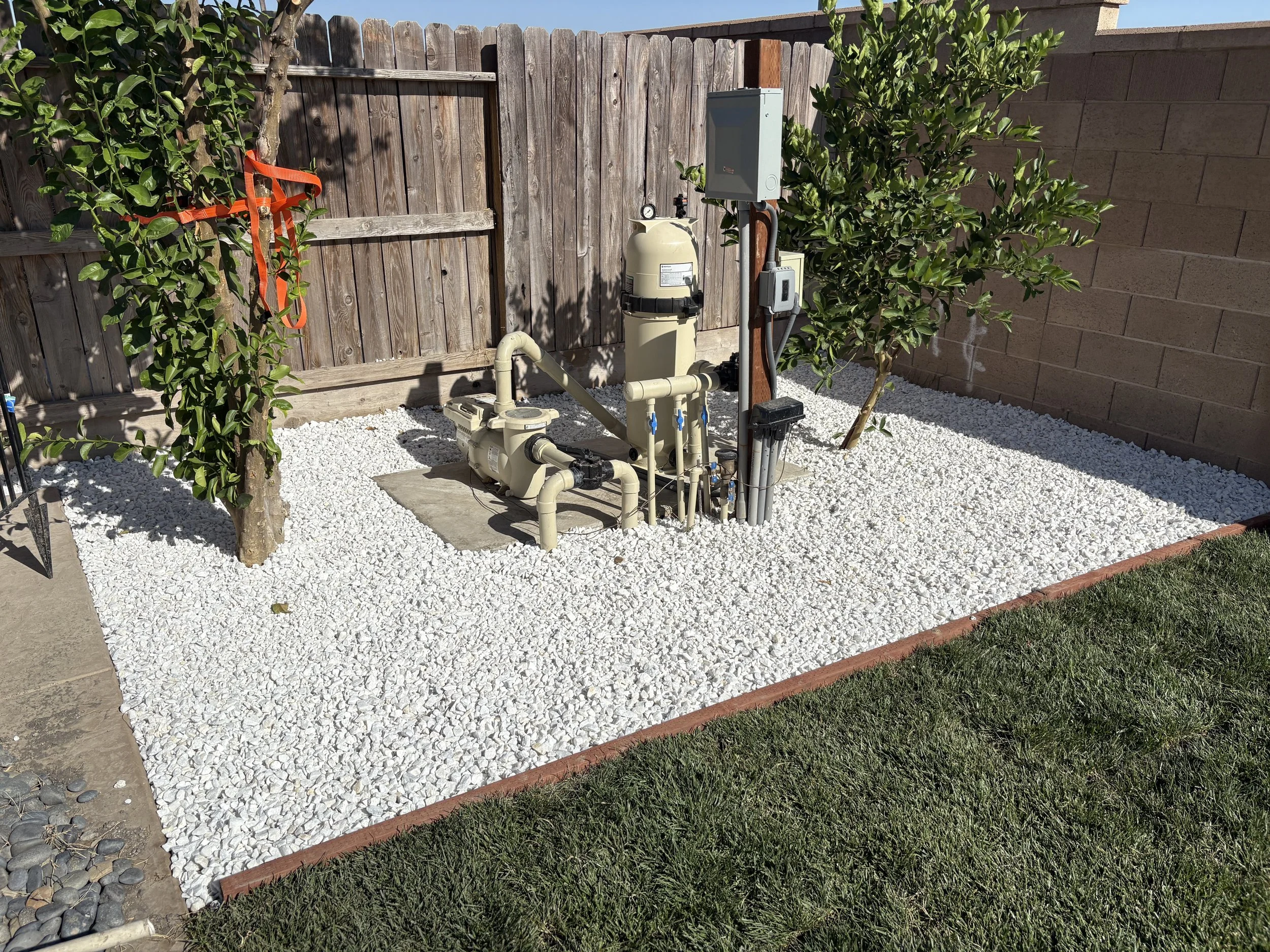 Backyard with pool equipment, including a filter, pump, and control box, surrounded by white gravel and bordered by grass and a wooden fence with a tree and bushes.