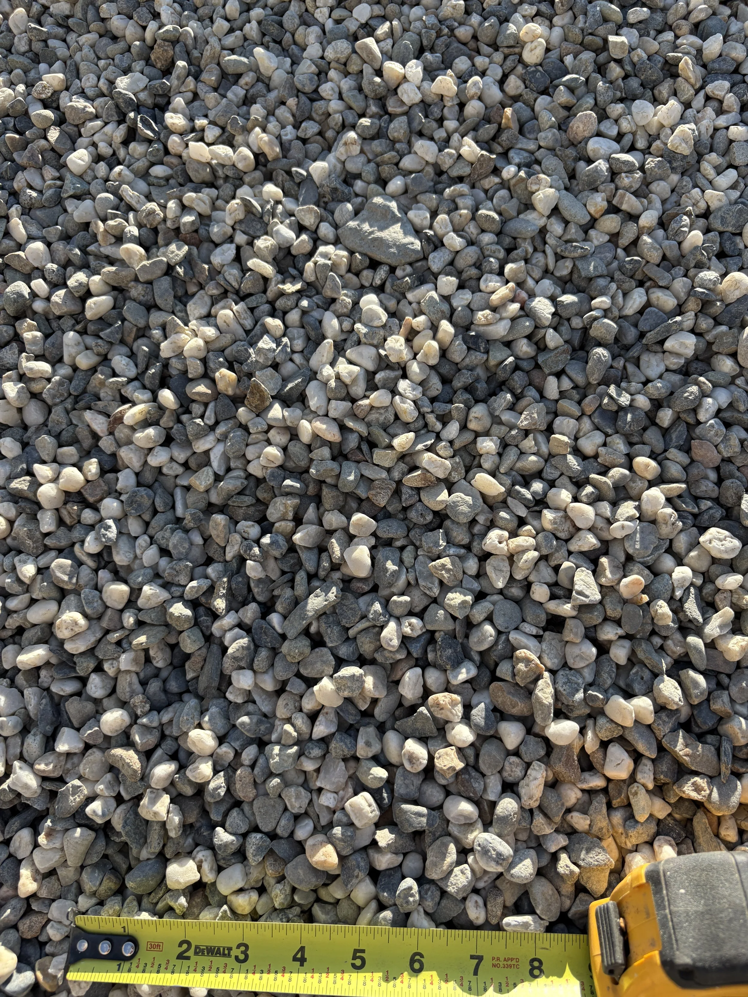 Close-up view of small, smooth gravel stones on the ground with a yellow measuring tape at the bottom showing approximately 8 inches and a portion of a yellow measuring device on the right side.