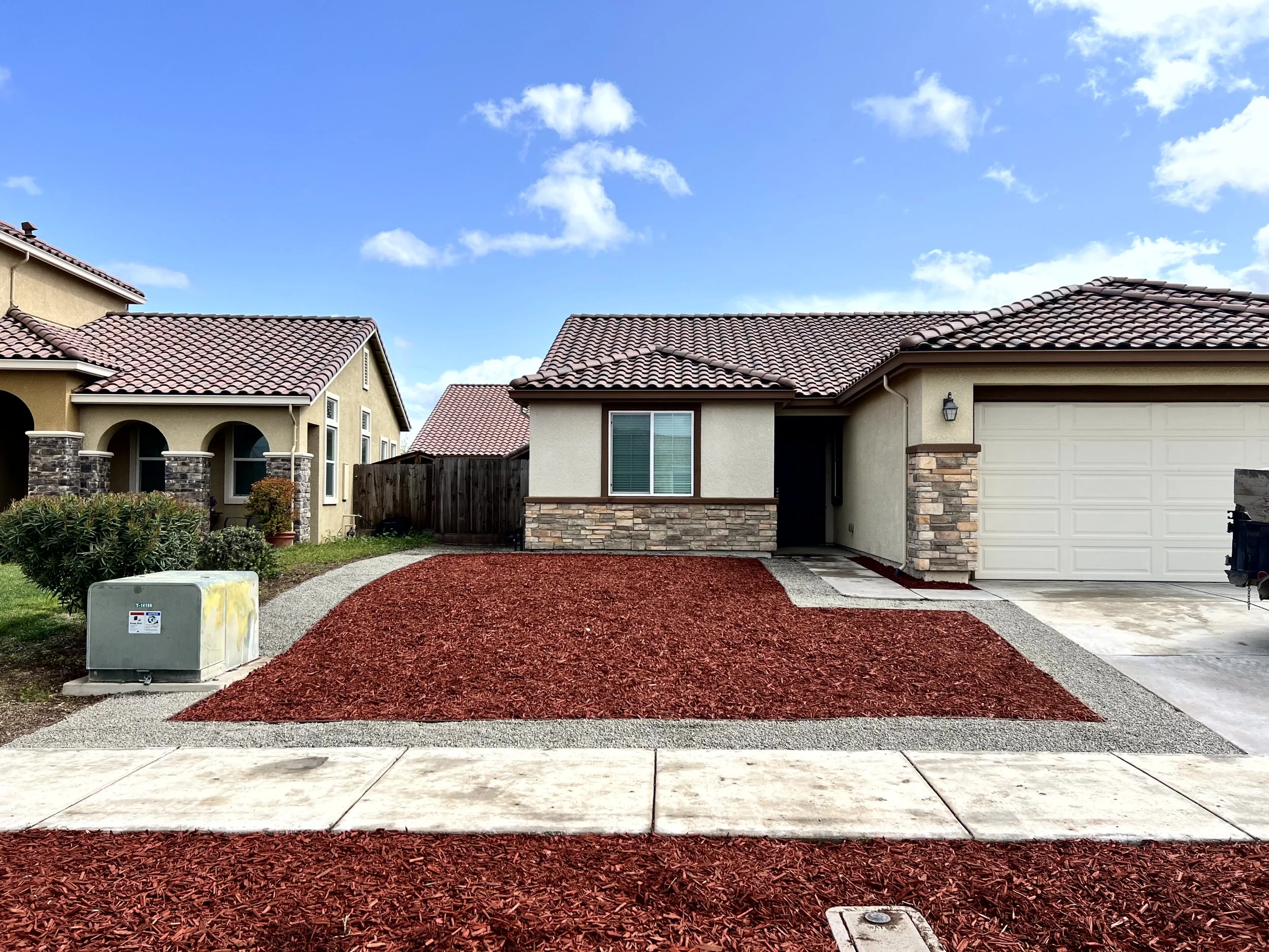 Front view of a single-story house with a garage, with a grass and red mulch yard, a concrete walkway, and neighboring houses under a blue sky with clouds.