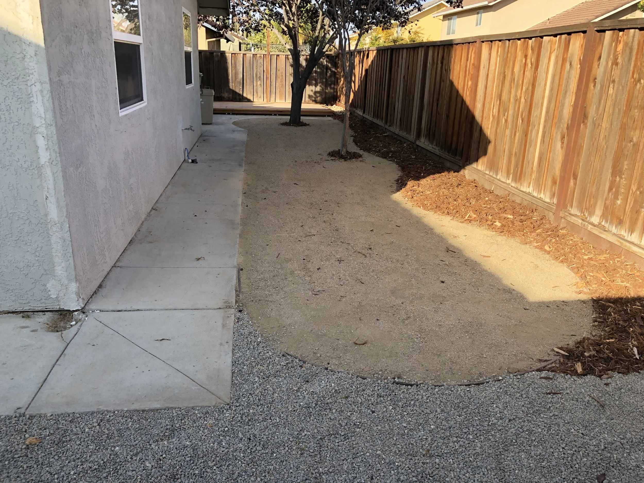 Backyard with a concrete patio and a small dirt patch adjacent to a house with beige stucco exterior, a tree, and a wooden fence.