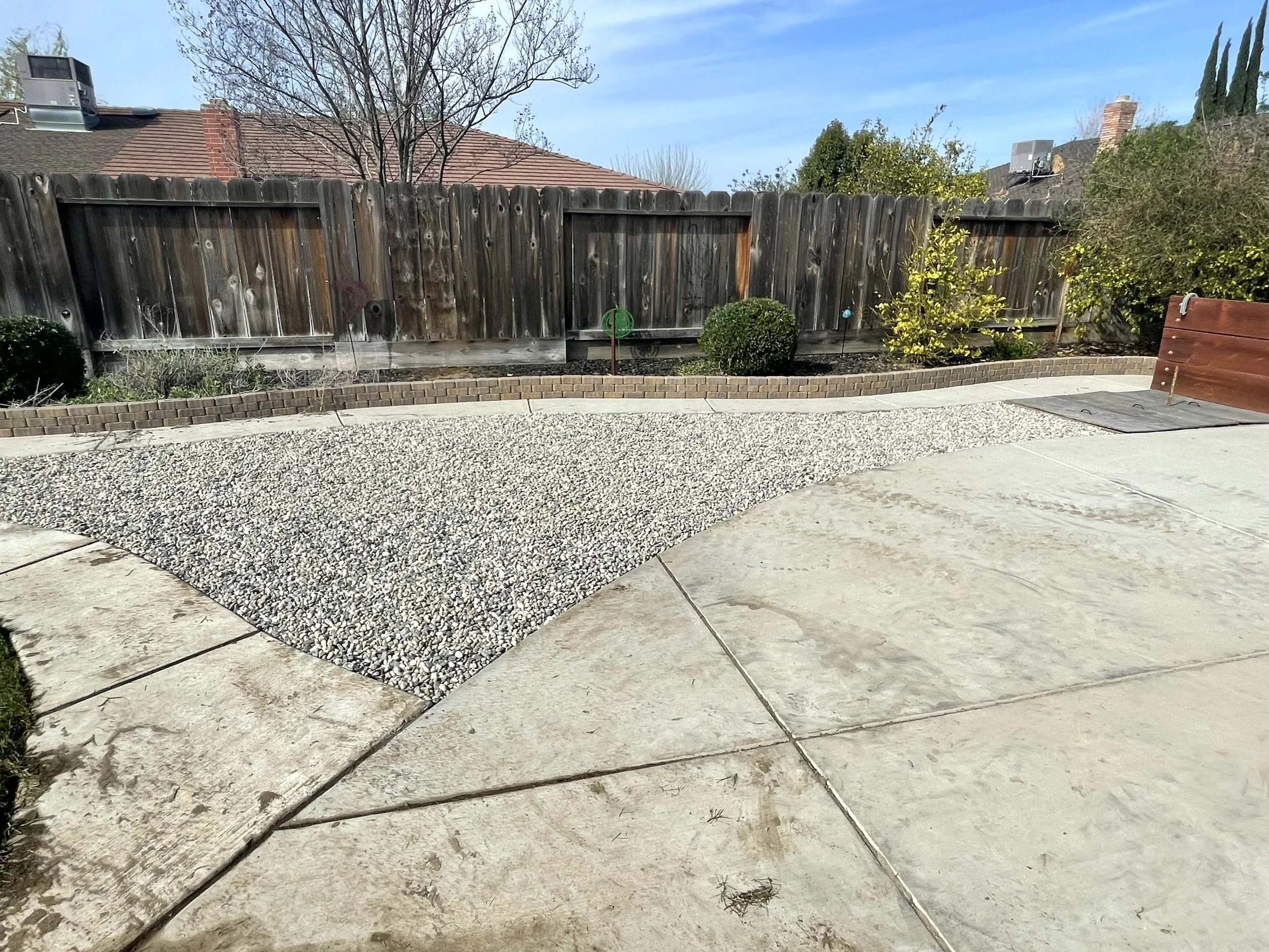 Backyard with a concrete patio, a gravel pathway, a wooden fence, and bushes.