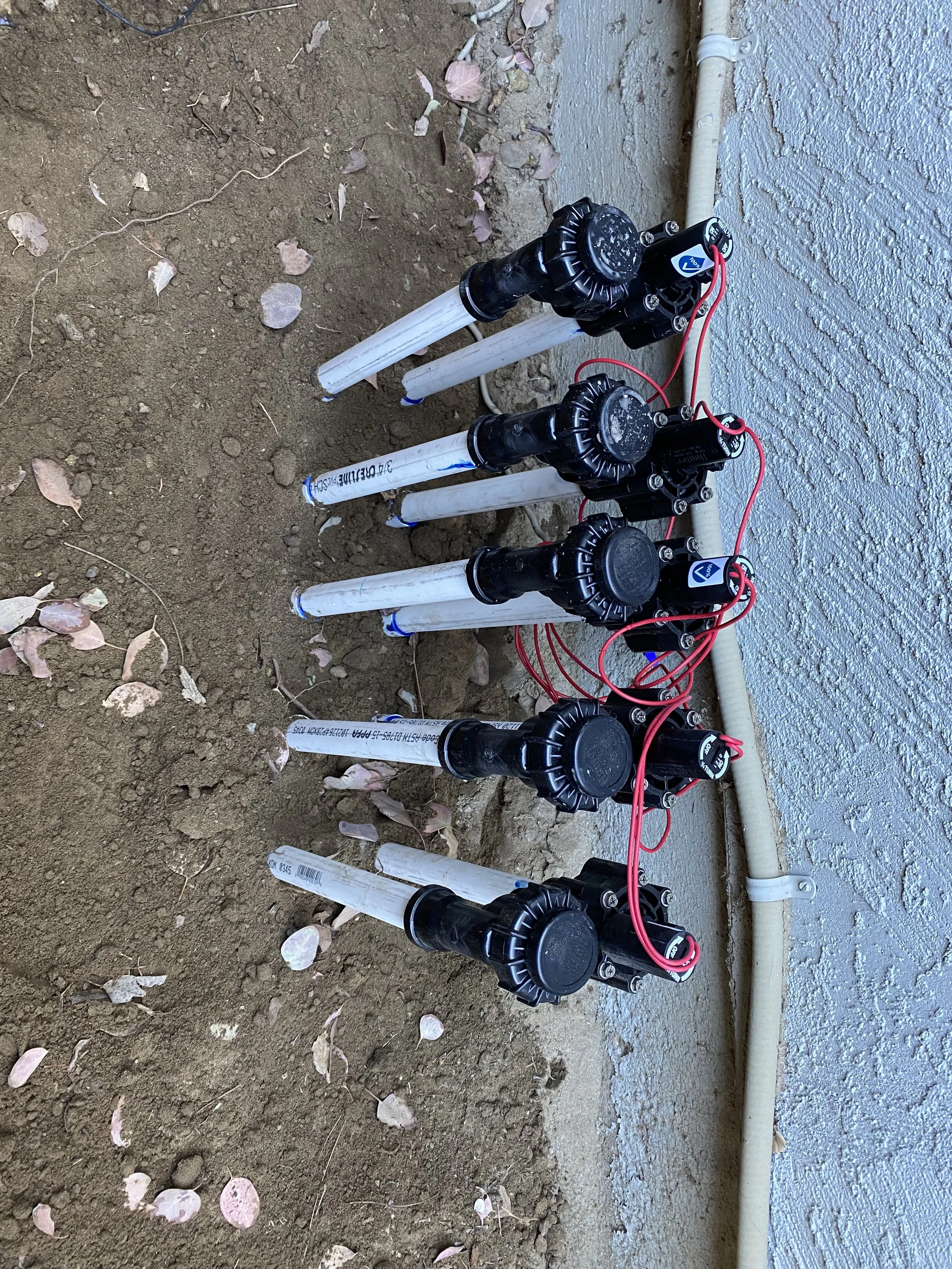 Water irrigation system with black valves and red wires on white PVC pipes installed in dirt next to a textured concrete wall with a pipe along the wall.
