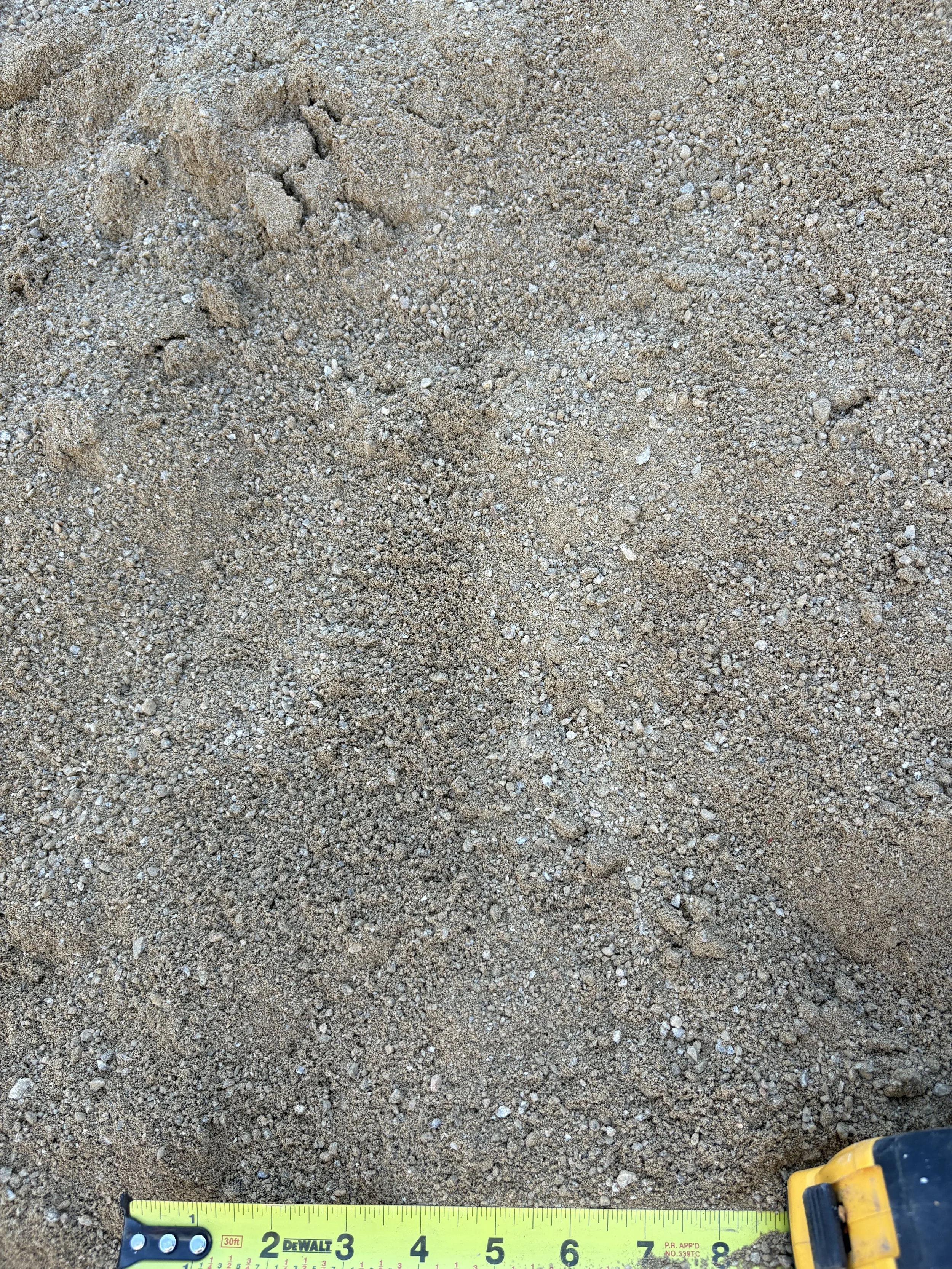 Close-up of sandy ground with small pebbles and a yellow measuring tape extending across the bottom of the image.