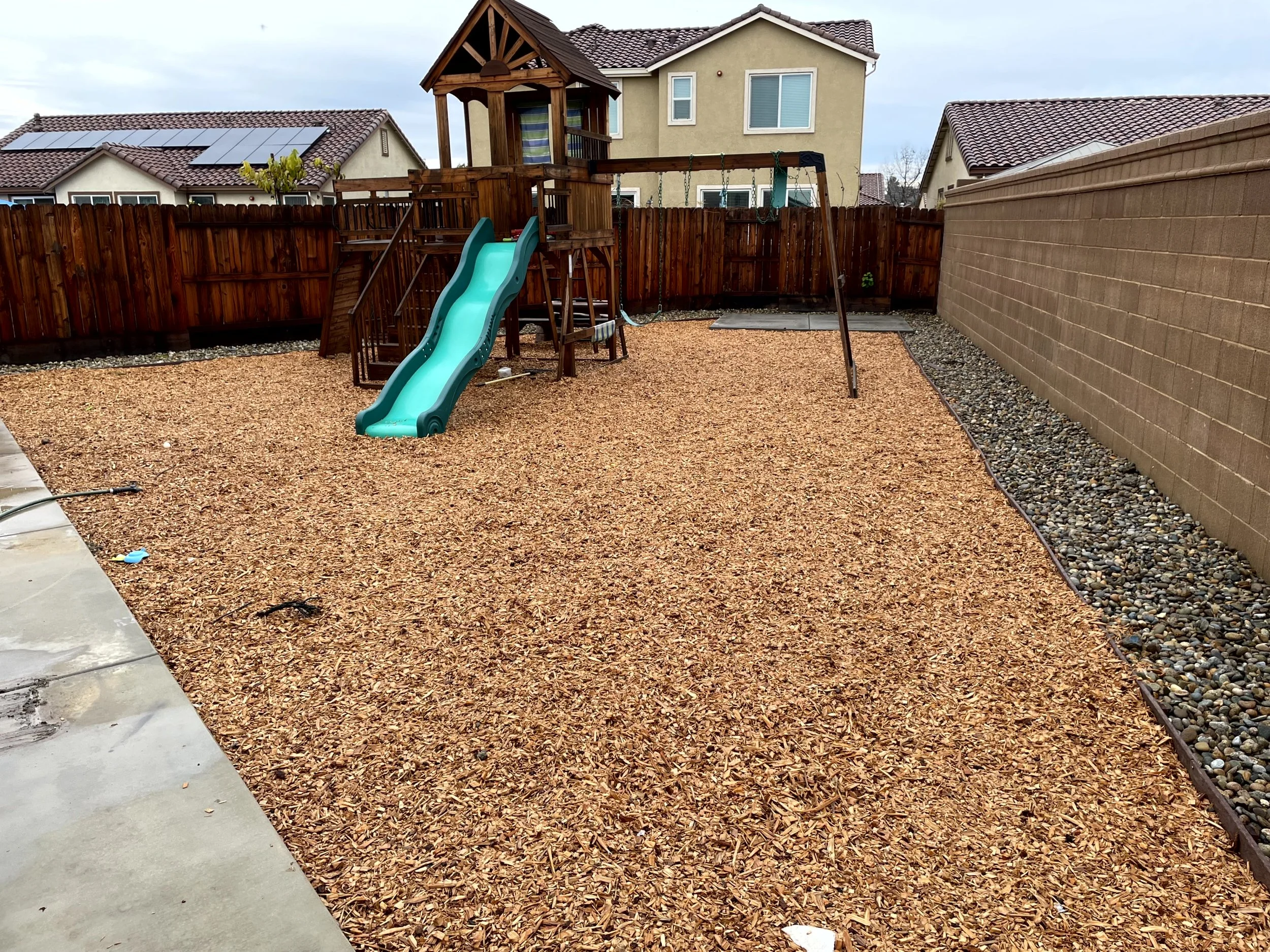 Backyard with wood chip ground covering, a wooden playset with slides and swings, surrounded by a wooden fence and a brick wall, with neighboring houses visible in the background.