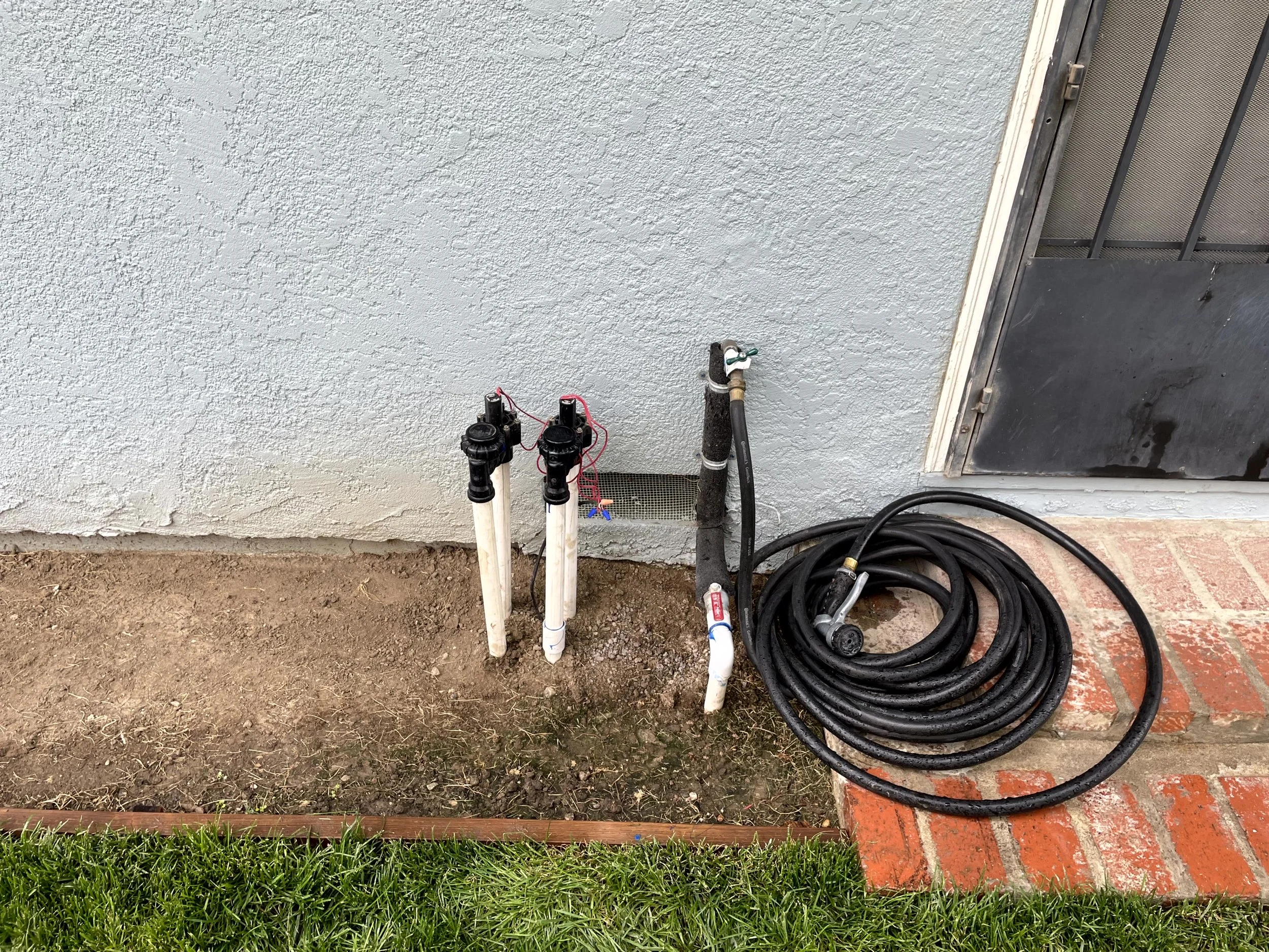 Garden hoses and sprinkler pipe fixtures attached to an exterior wall of a house, with a coiled hose lying on the brick step next to the door.