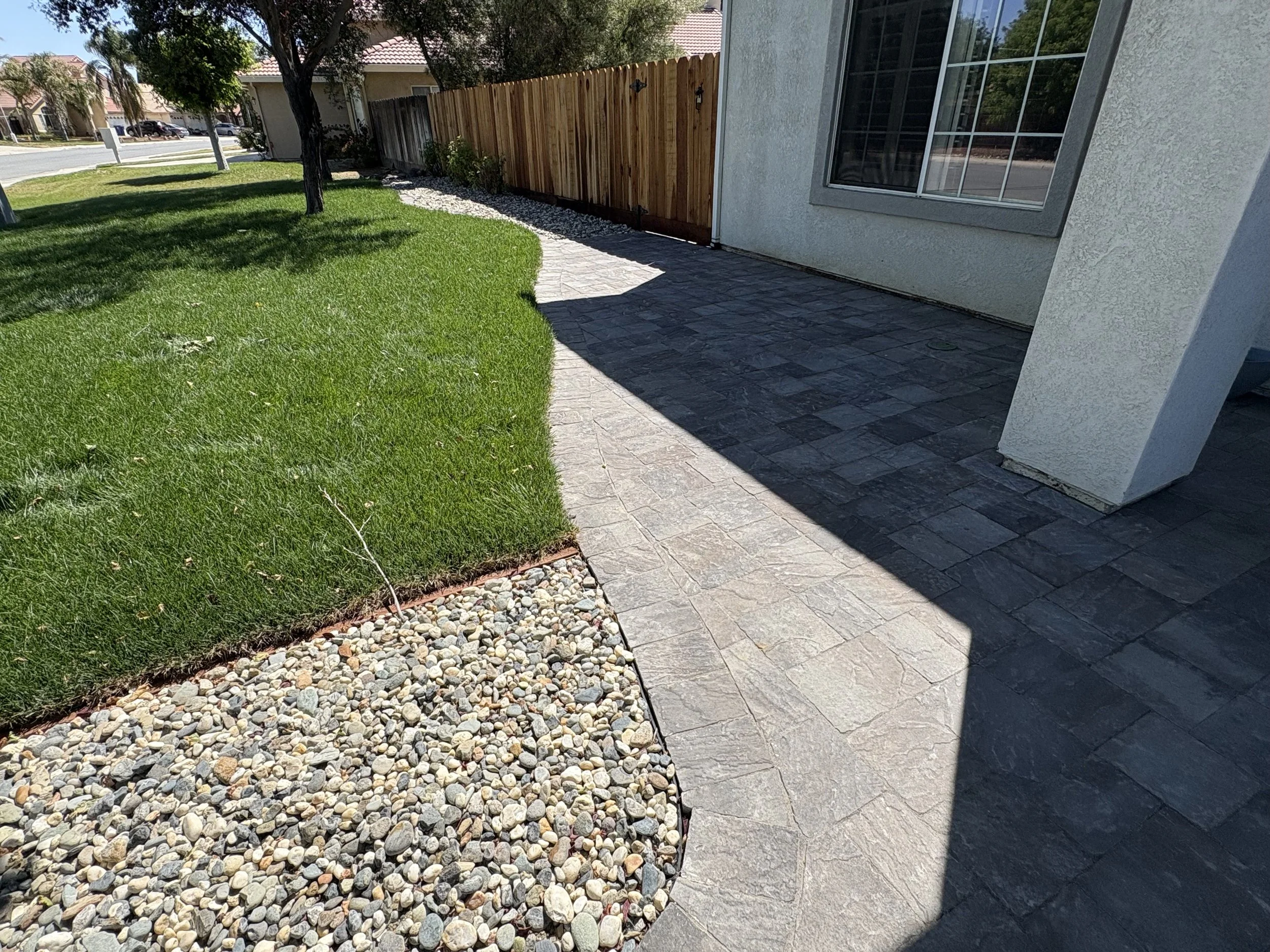 Backyard with a patch of green grass, decorative rocks, and a paved patio area next to a house with large windows.