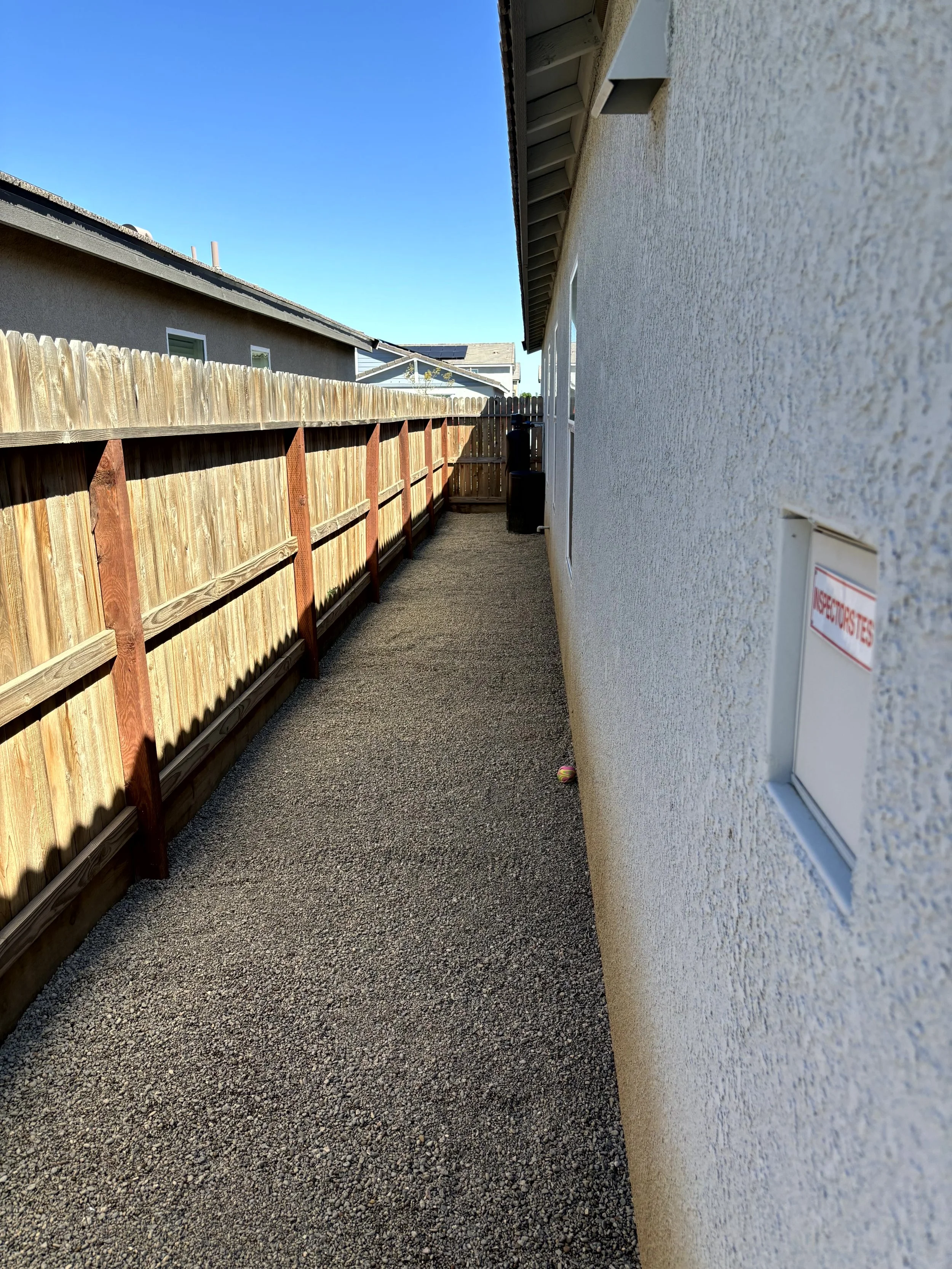 Narrow backyard side yard with gravel pathway, wooden fence on the left, white stucco house wall on the right, and a small ball on the ground near the house wall, with a clear blue sky above.