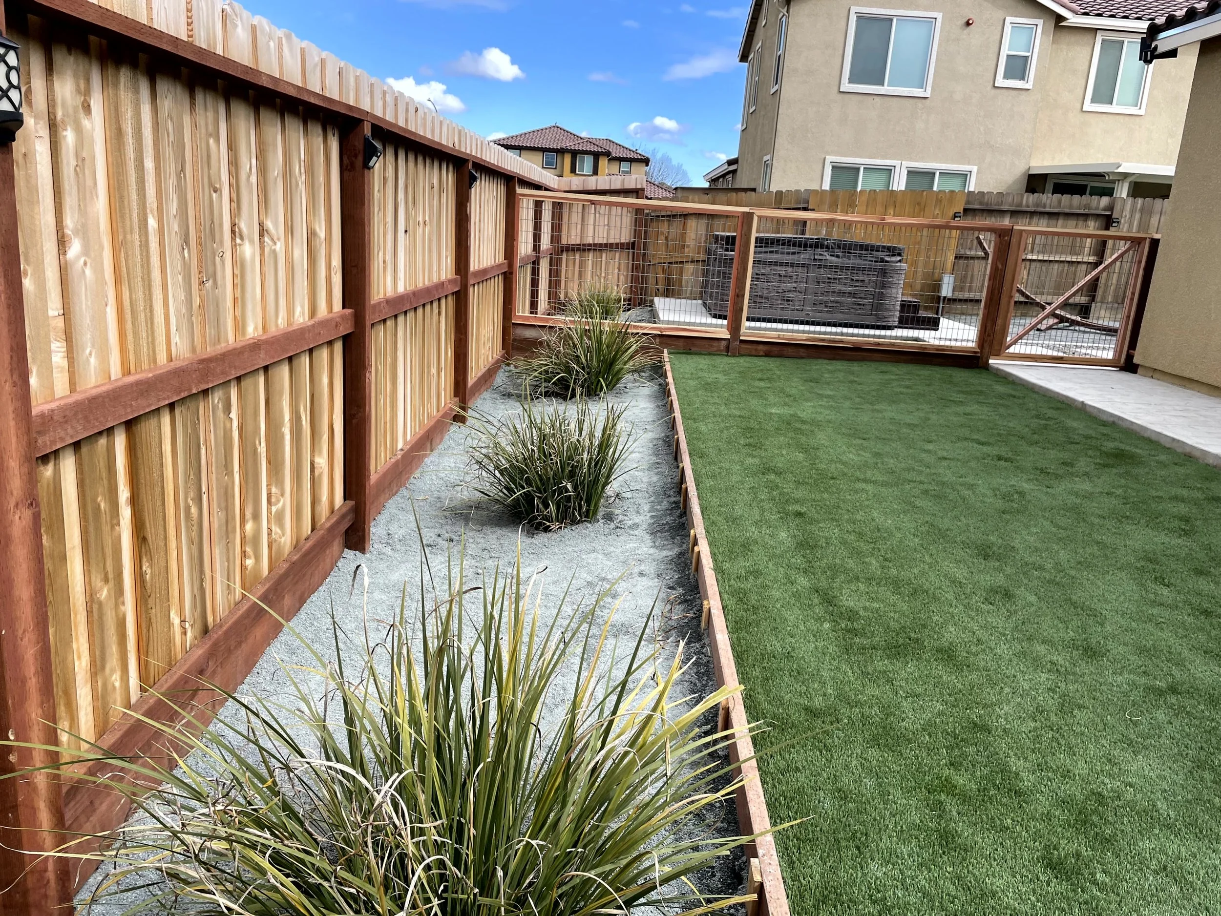 A backyard with a wooden fence, artificial grass, and a garden bed with ornamental grasses, under a partly cloudy sky.