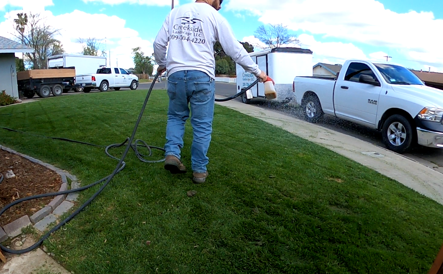 A person watering a lawn with a hose connected to a spray nozzle, near a sidewalk and a parked white pickup truck.