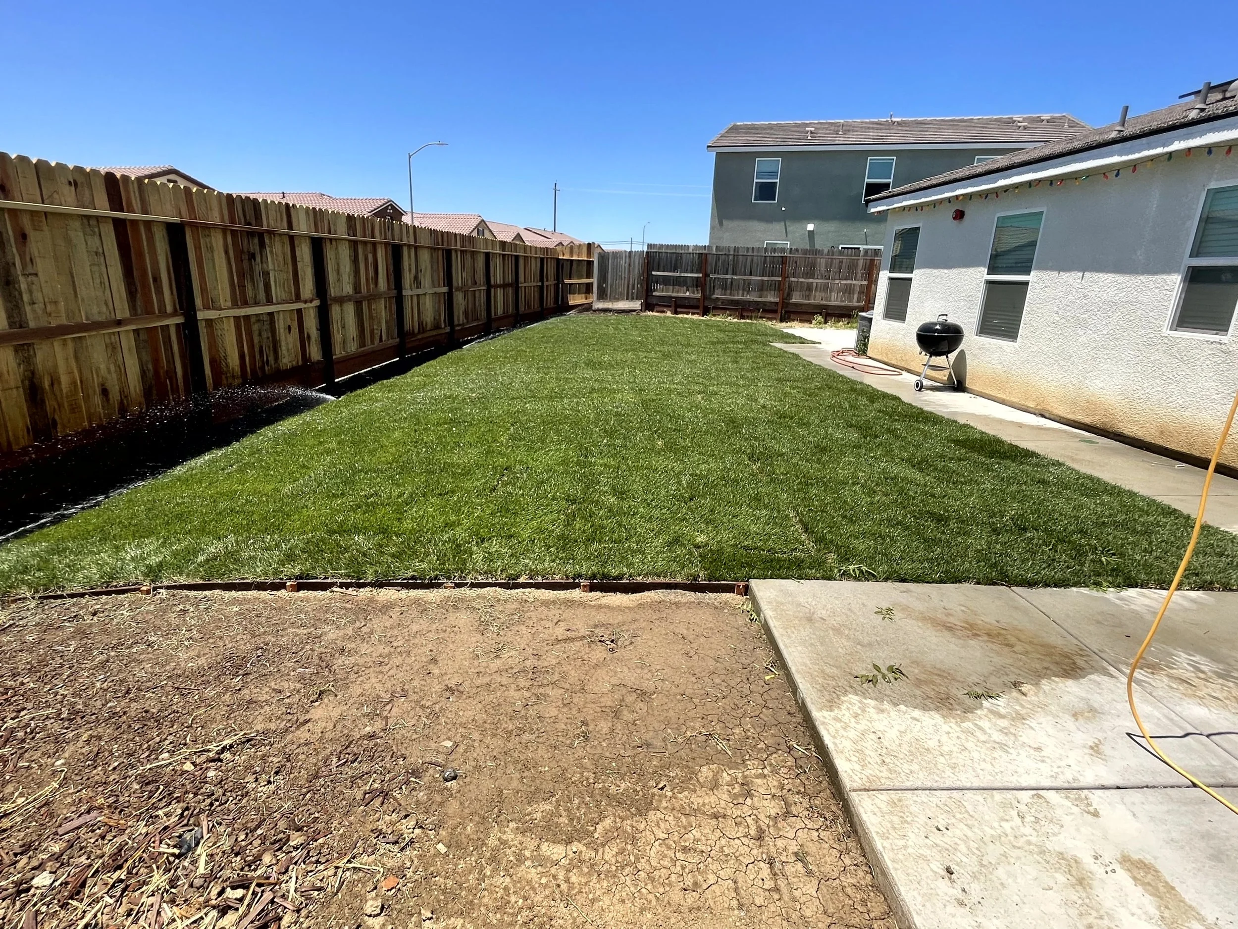 View of a backyard with a freshly mowed green lawn, surrounded by wooden fences, with a patio area and a barbecue grill near a white house, under a clear blue sky.