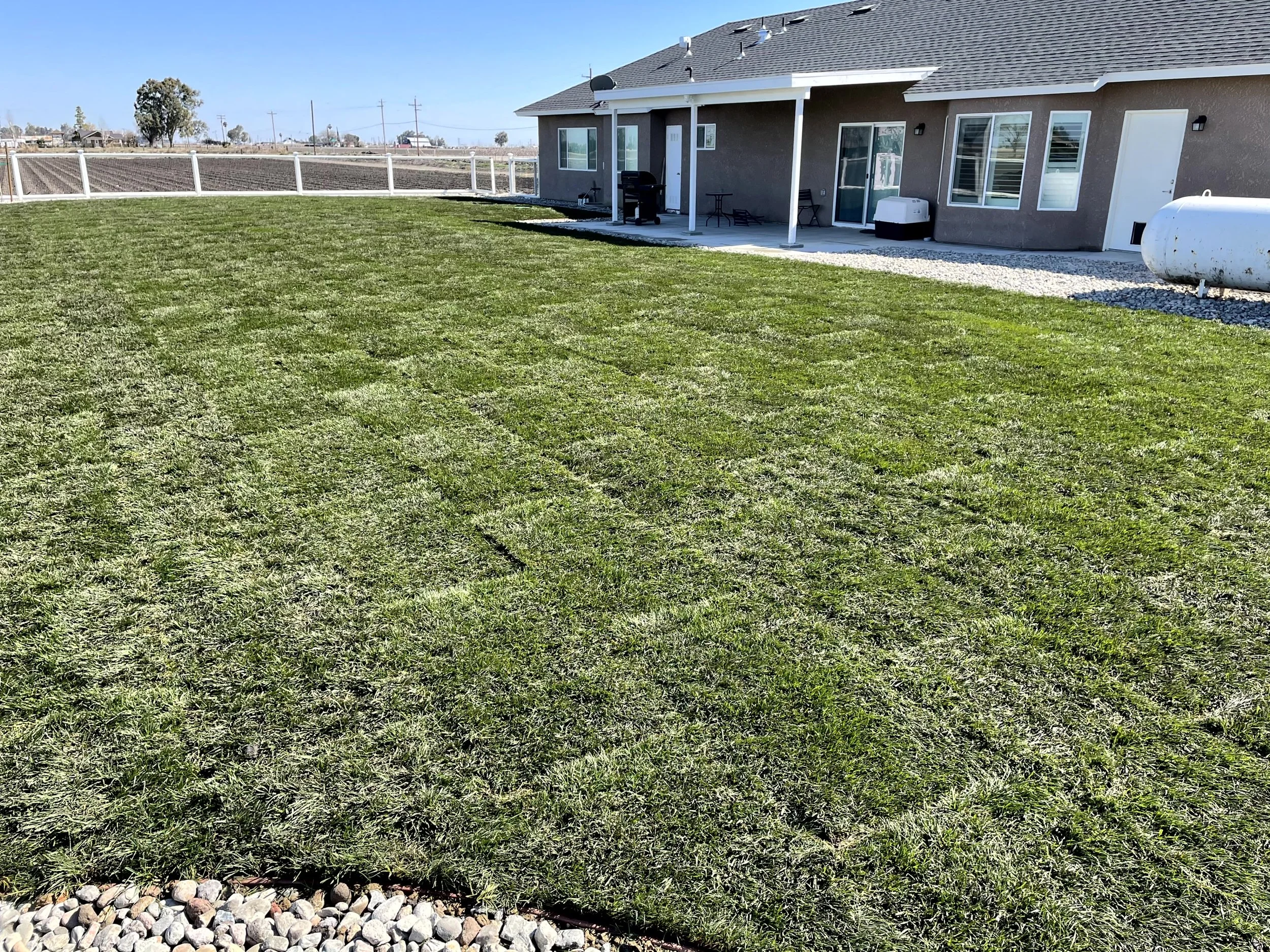 Backyard with freshly laid green grass, a gray house with multiple windows and a sliding door, patio furniture, a propane tank, and a gravel path, enclosed by a white fence.