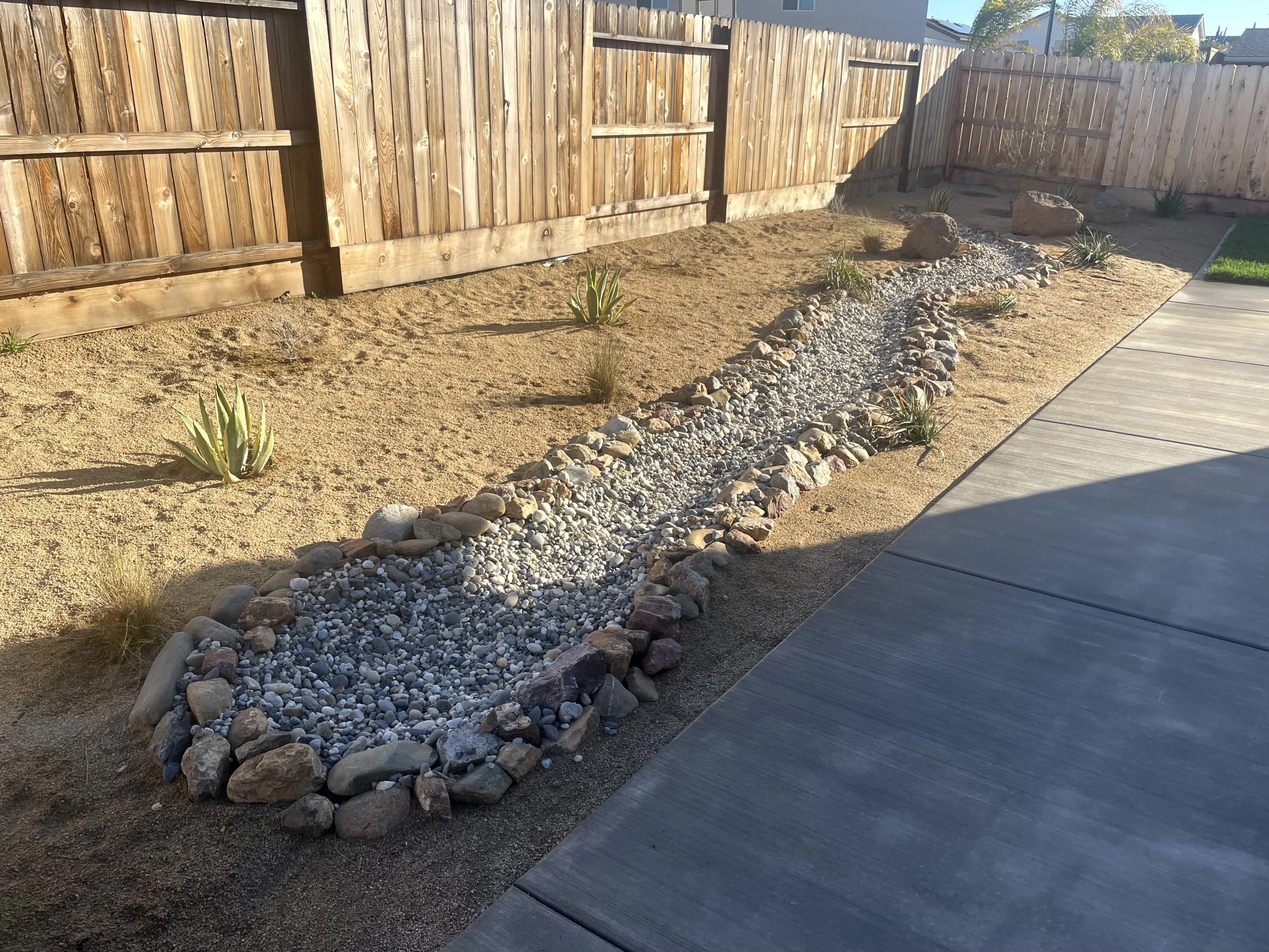 Backyard landscape with a gravel stream, desert plants, potted plants, a wooden fence, and a concrete patio.