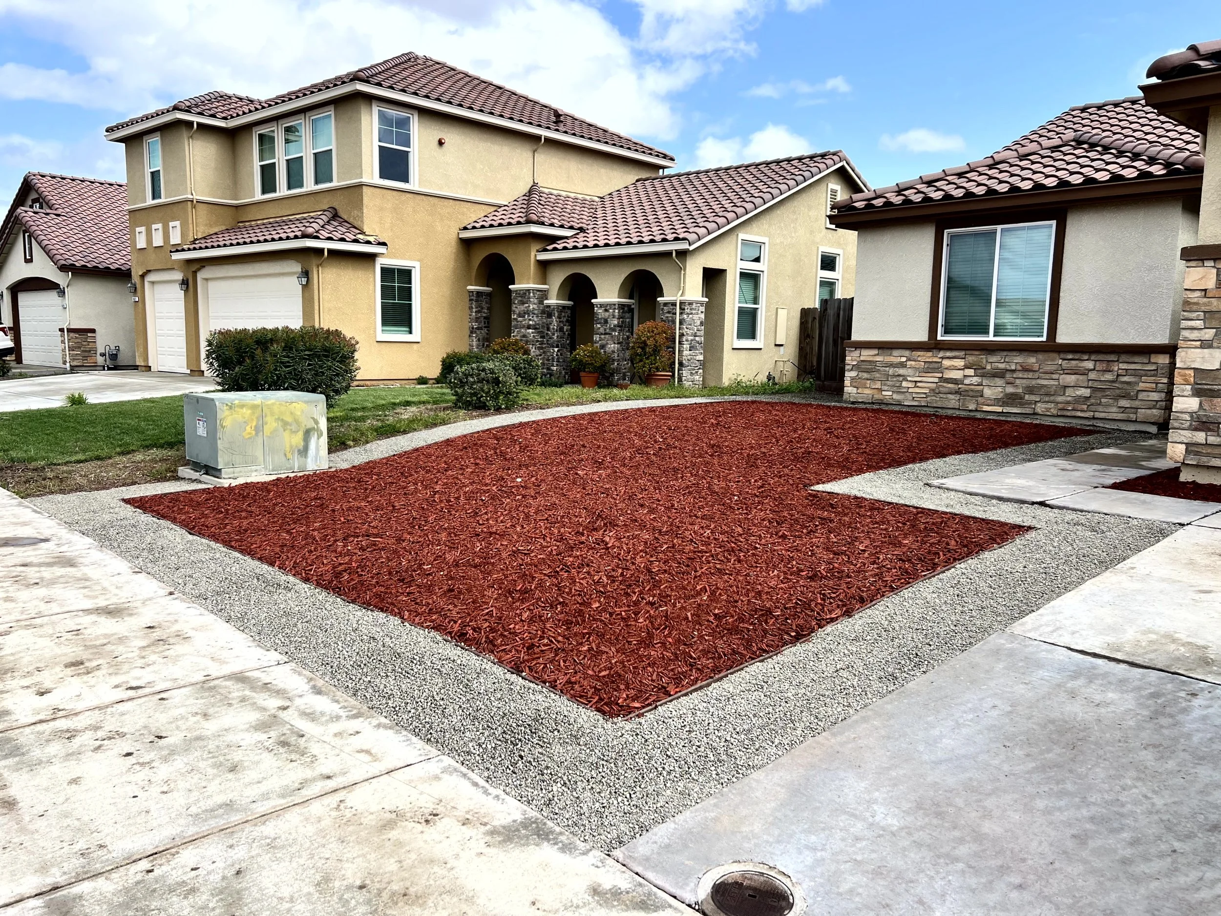 A suburban house with a front yard featuring decorative red mulch bordered by gravel, with neighboring houses and a blue sky with clouds.