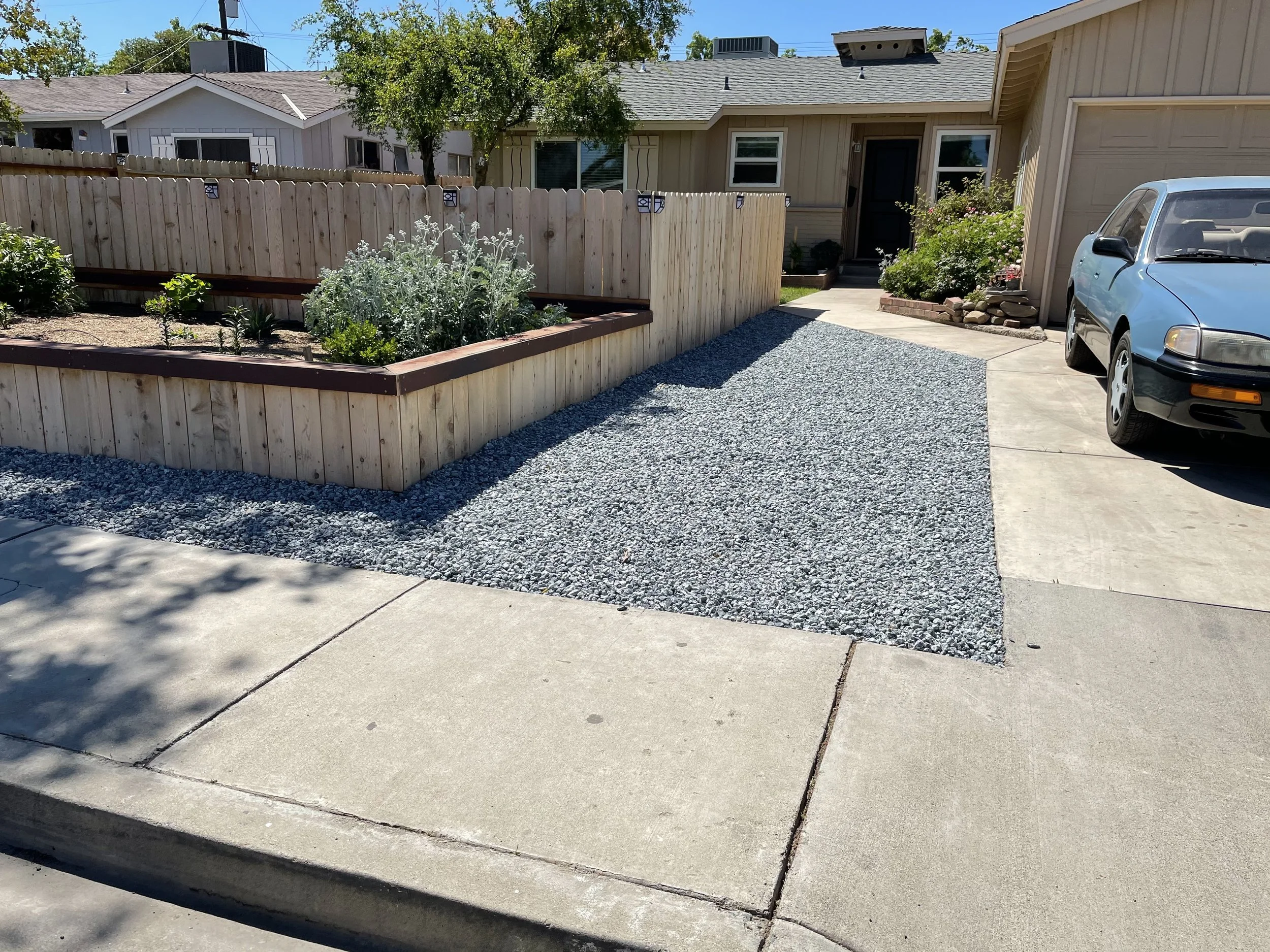 A residential driveway with a section of gravel, a parked blue car, and a garden bed with plants and flowers enclosed by a wooden fence.