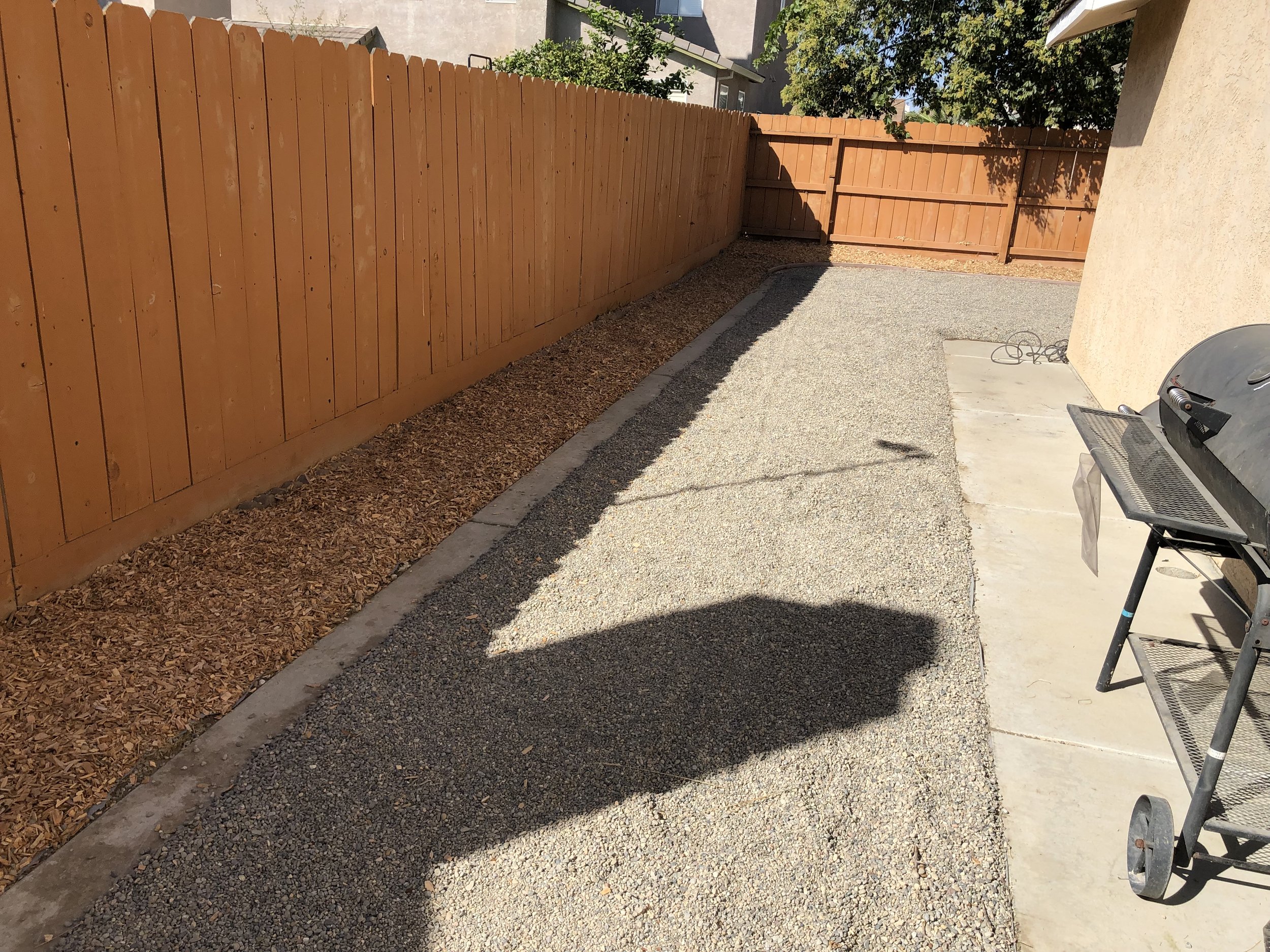 A small backyard patio with a gravel ground, bordered on one side by a wooden fence and on the other by a house wall with a grill and a small cart nearby. There is a shadow cast on the gravel from an object out of view.