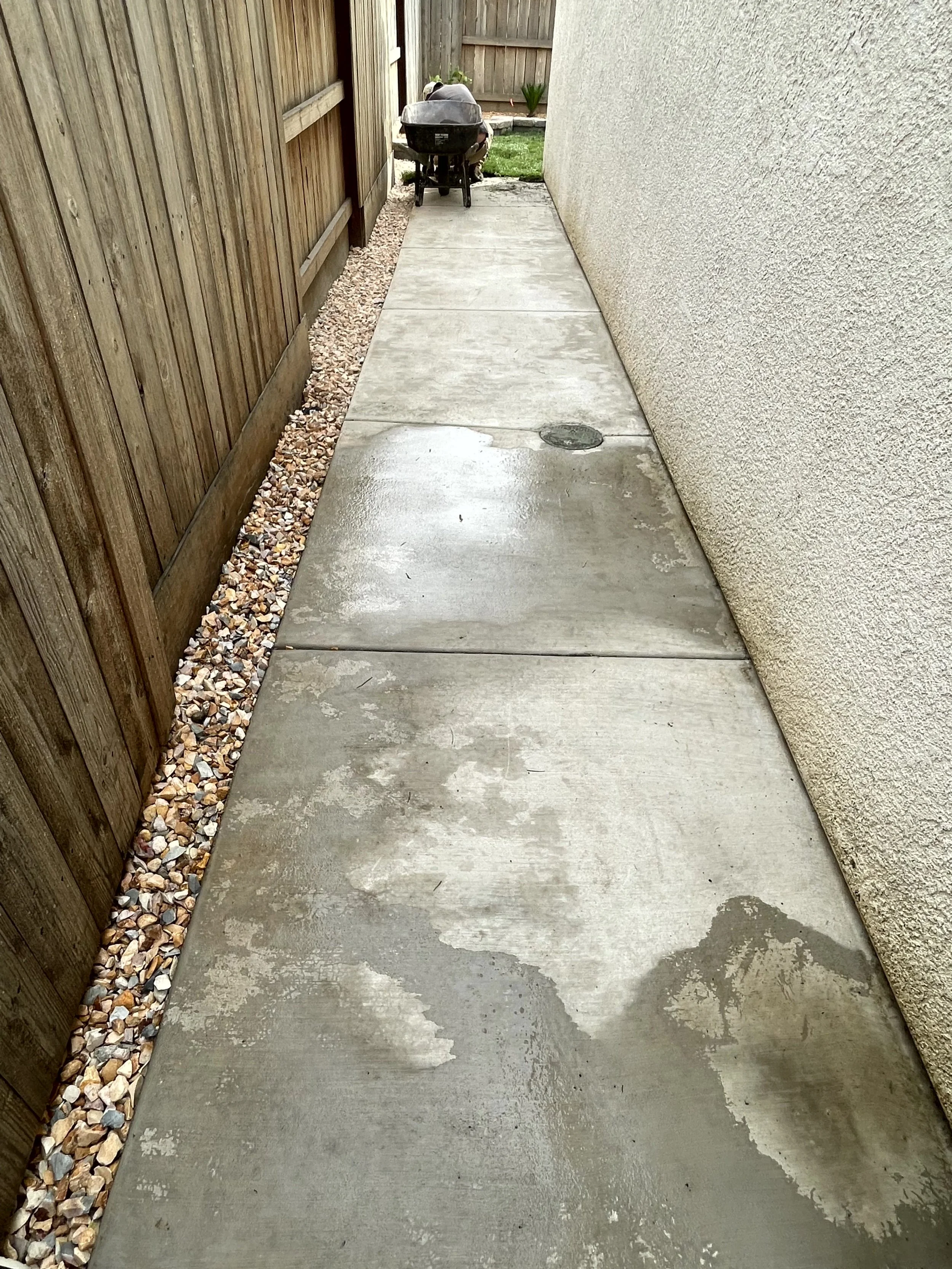 Concrete walkway with water stains, adjacent to a wooden fence on the left and a textured beige wall on the right, leading to a small yard with a grill and grass. 