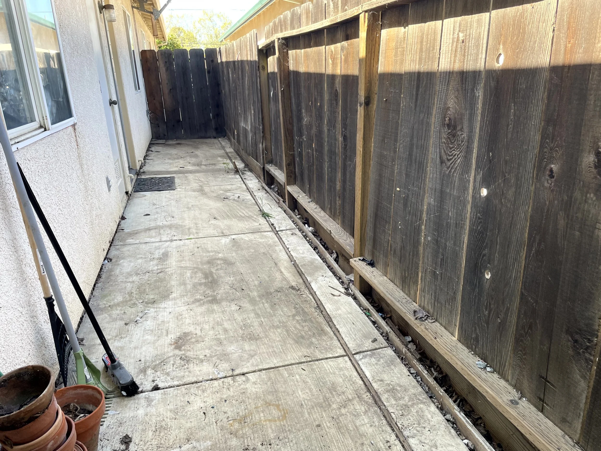 Side yard with concrete patio, black wooden fence partially constructed, and gardening tools leaning against the house wall.