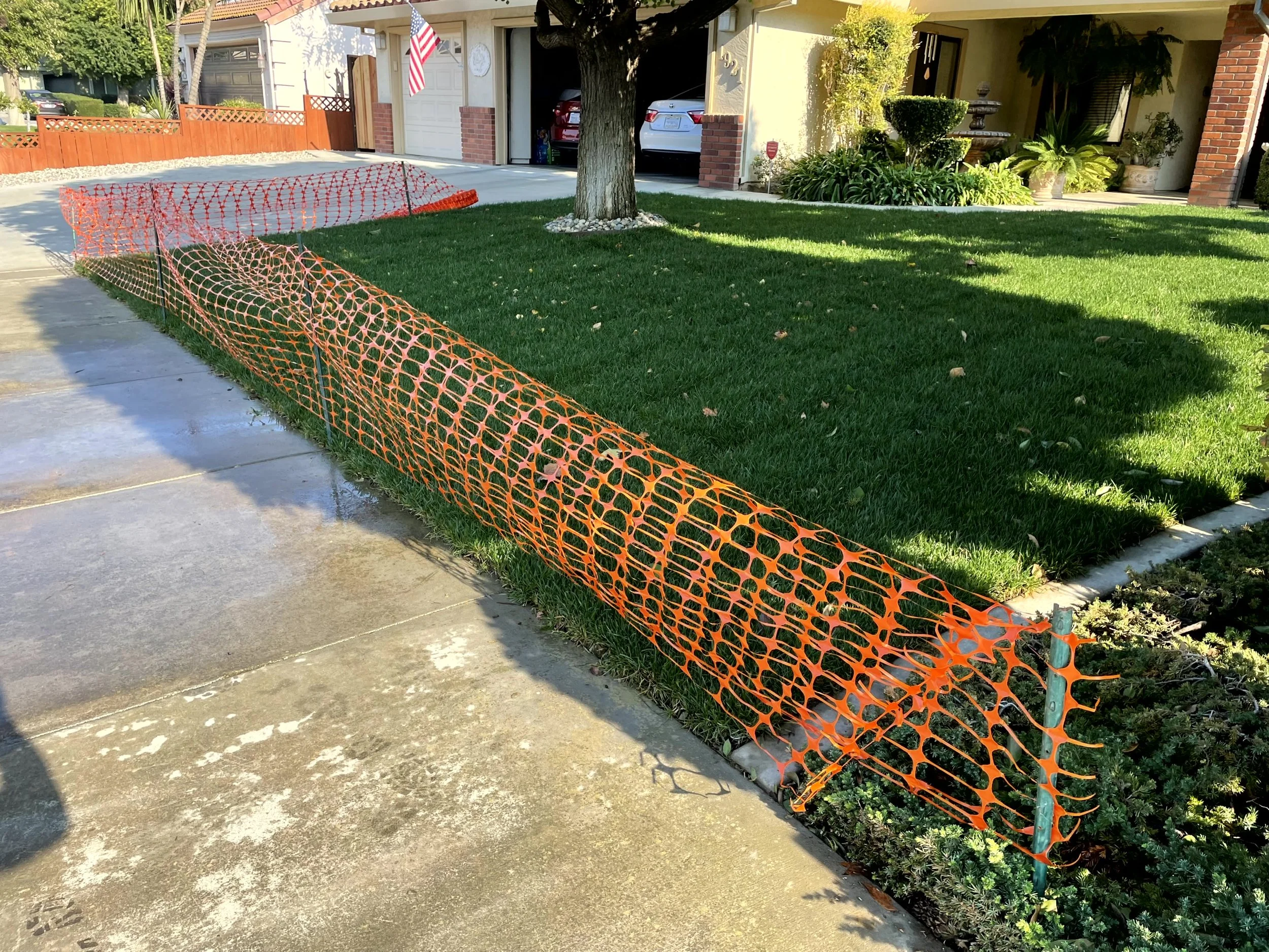 Orange plastic safety fence around a lawn and garden area in front of a house, with a sidewalk in the foreground and cars parked in the driveway.
