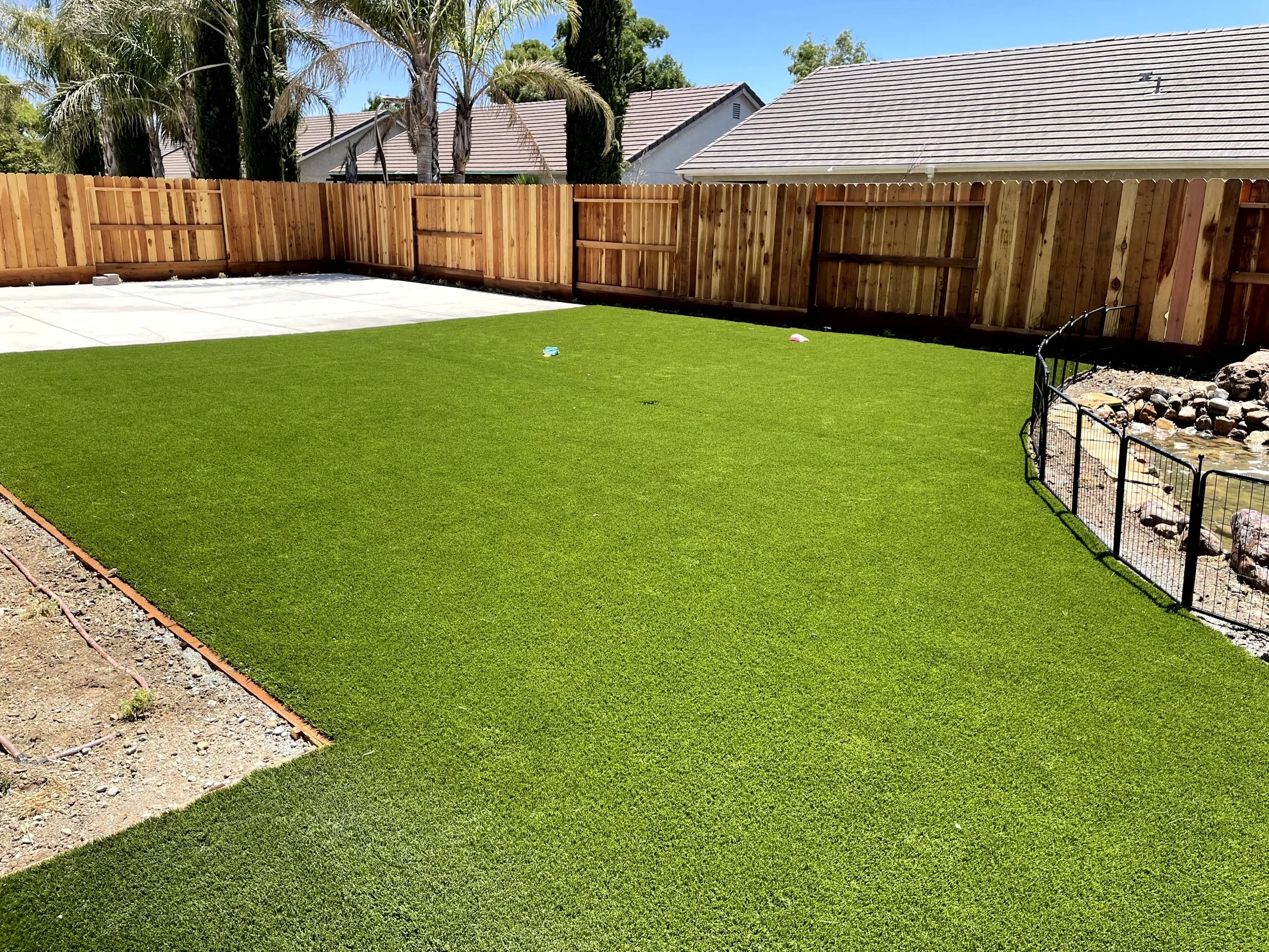 A backyard with a lush, green artificial grass lawn, a fenced pond with rocks, a wooden privacy fence, and a concrete patio area in the background.