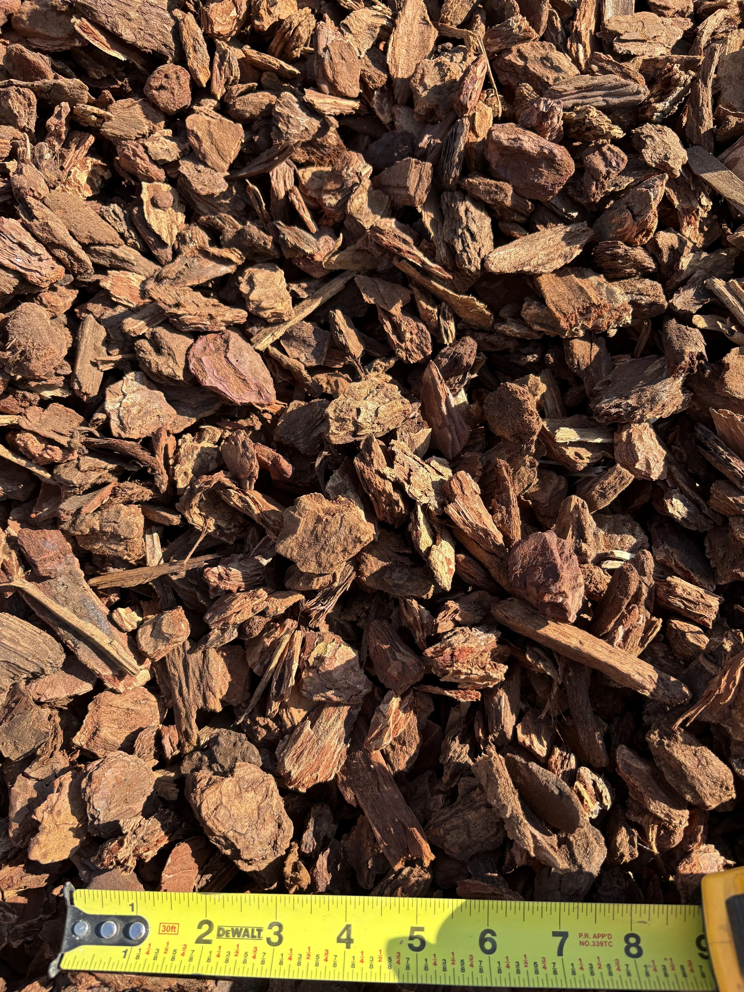 Close-up of a pile of brown bark mulch, with a yellow measuring tape at the bottom showing inches.