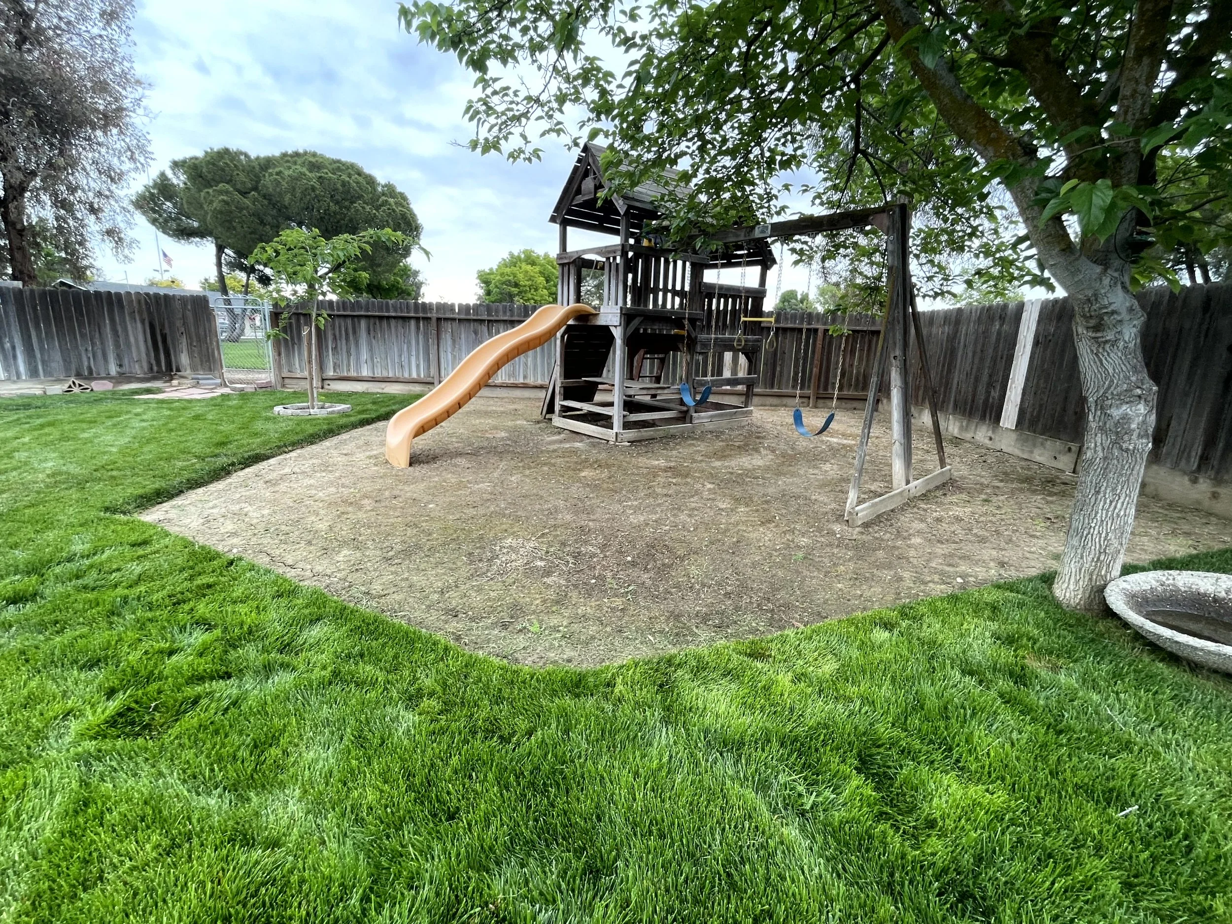Backyard playground with a slide, swings, and a wooden play structure surrounded by a wooden fence, green grass, trees, and cloudy sky.