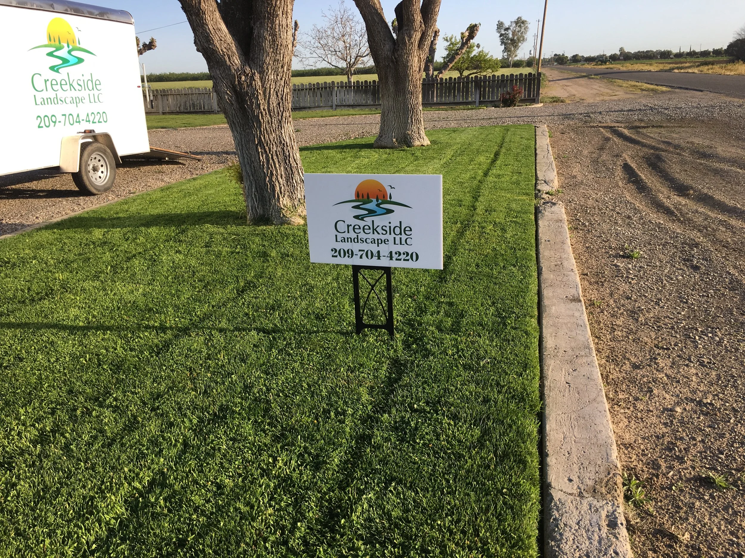 A sign for Creekside Landscape LLC with a phone number, placed on a small patch of green grass next to a larger tree and a gravel driveway in a rural area.