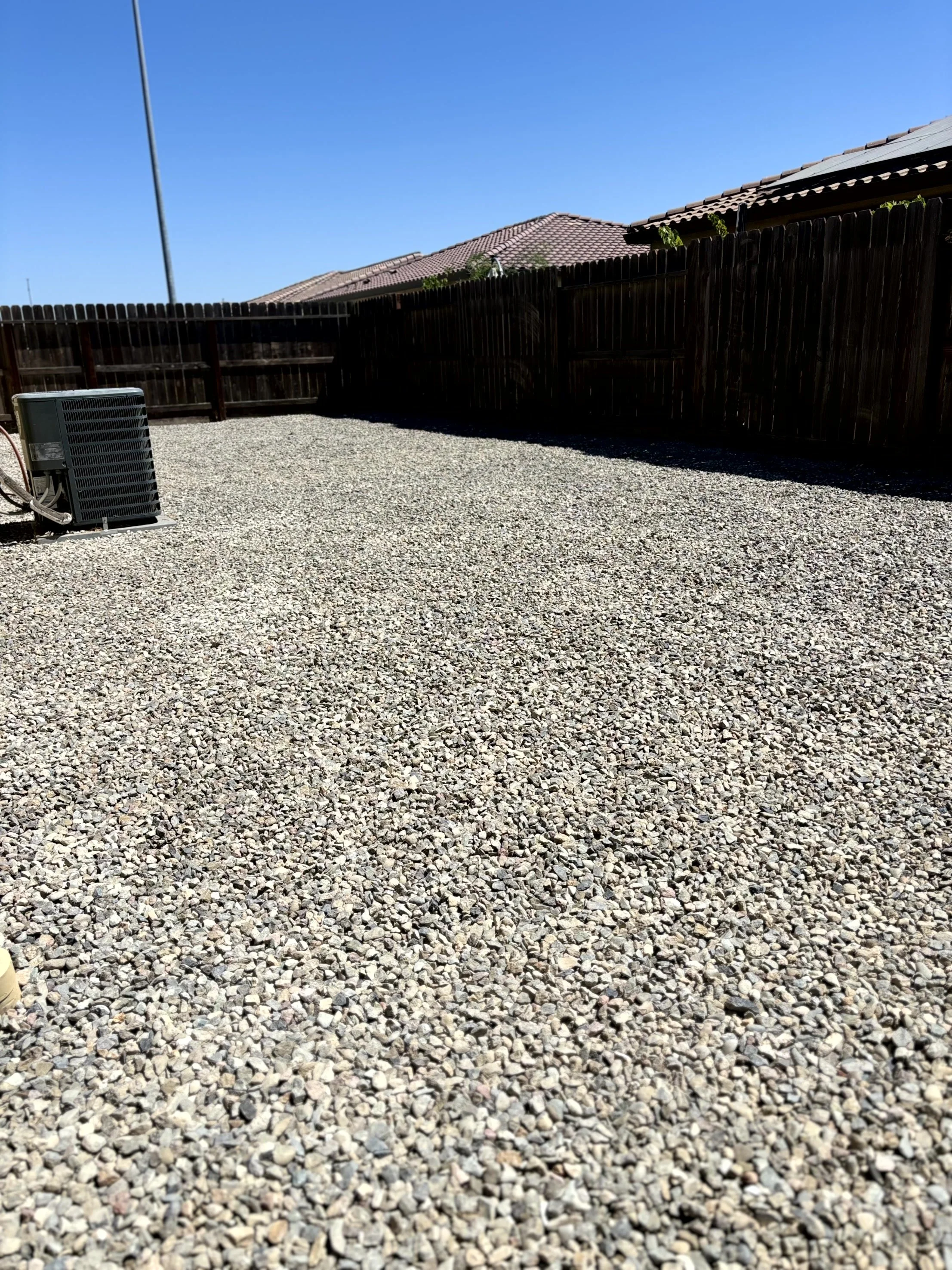 Backyard with gravel ground, wooden fence, and air conditioning unit, under clear blue sky.