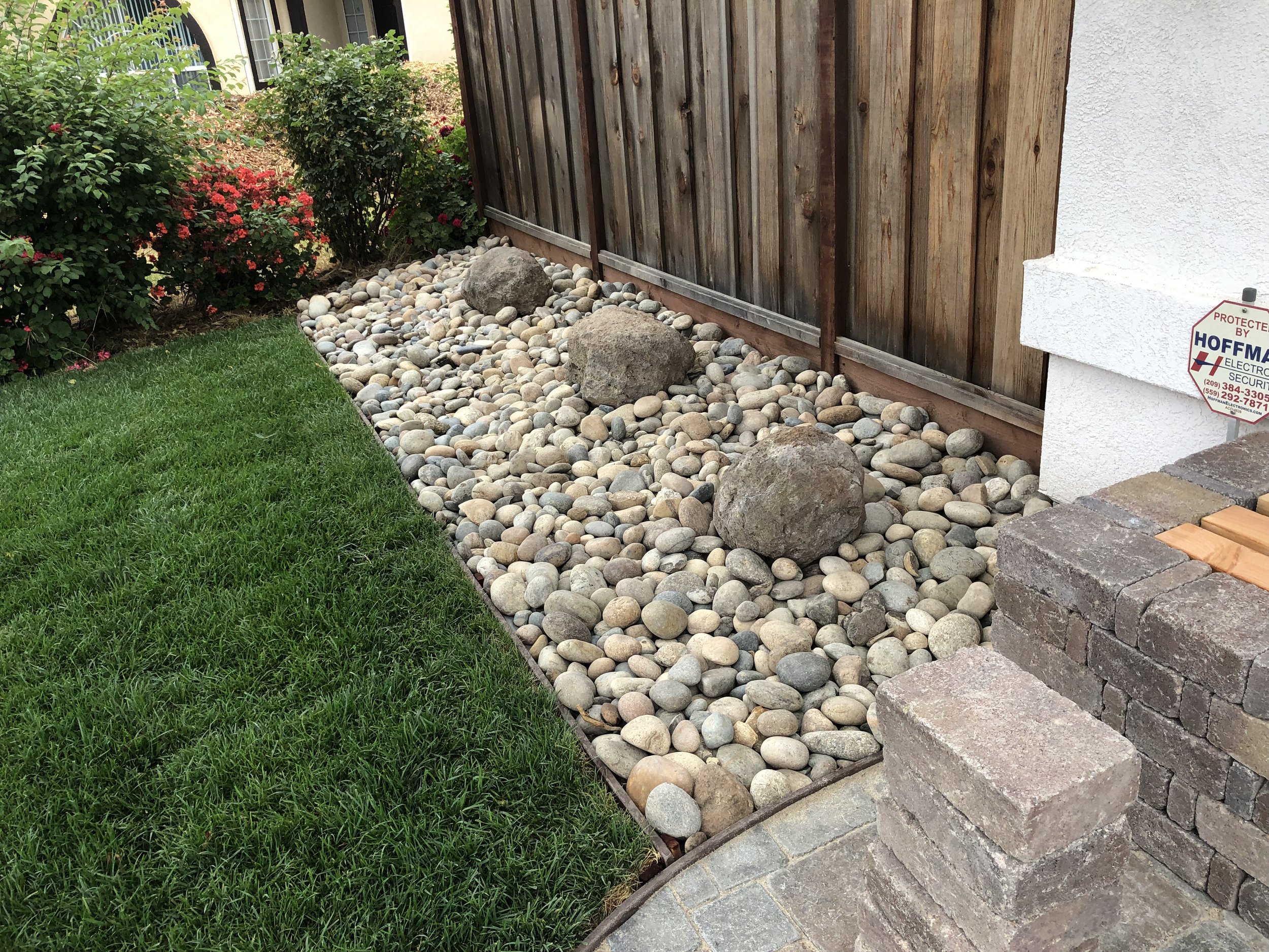 A landscaped yard with a grass lawn, a flower bed with red flowers, and a section with smooth rocks and large stones separated by a border, next to a wooden fence and a white wall with a security sign.