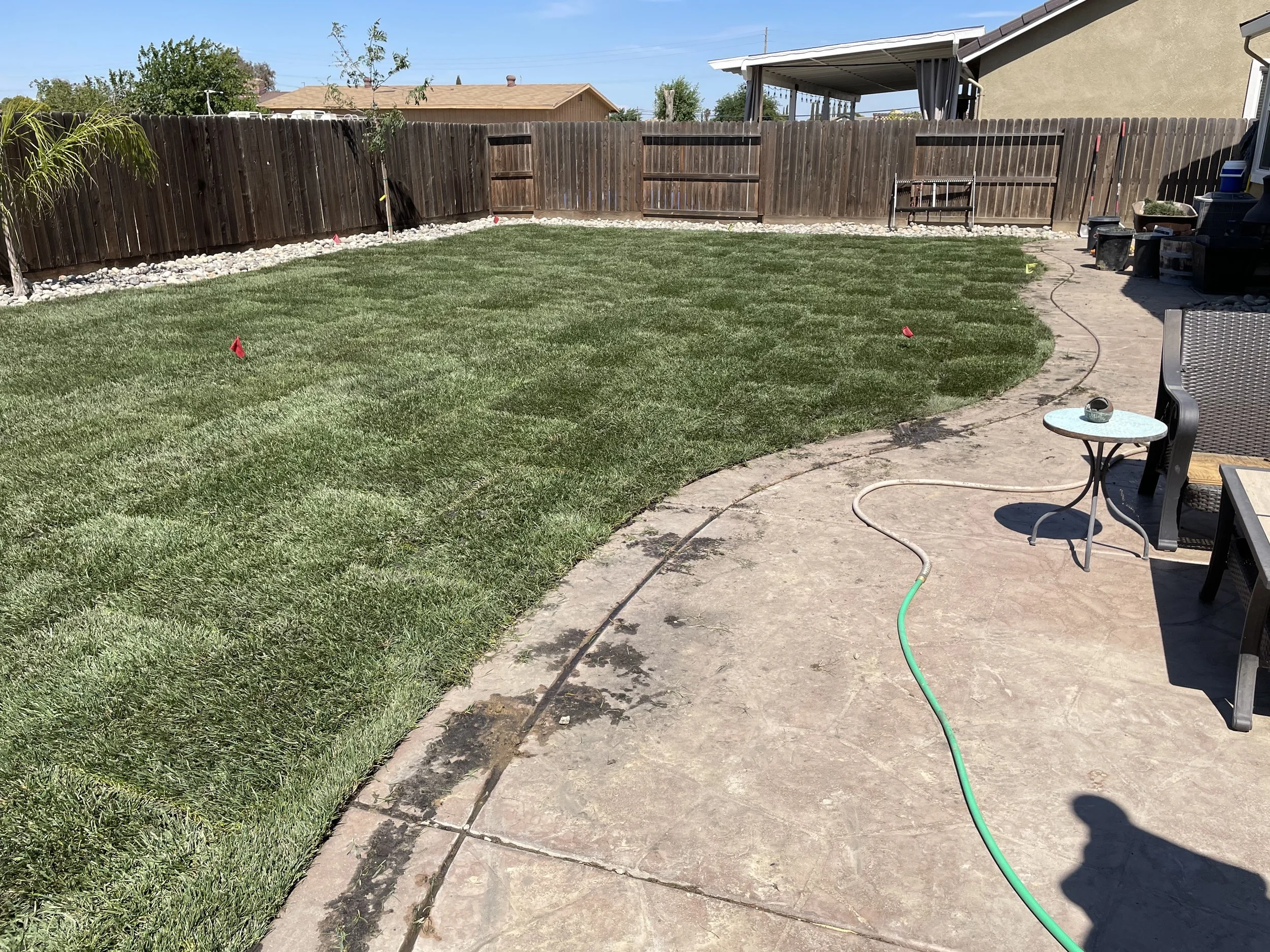 A backyard with freshly laid sod grass, a stone patio with outdoor furniture, a garden hose, and a wooden fence around the yard.