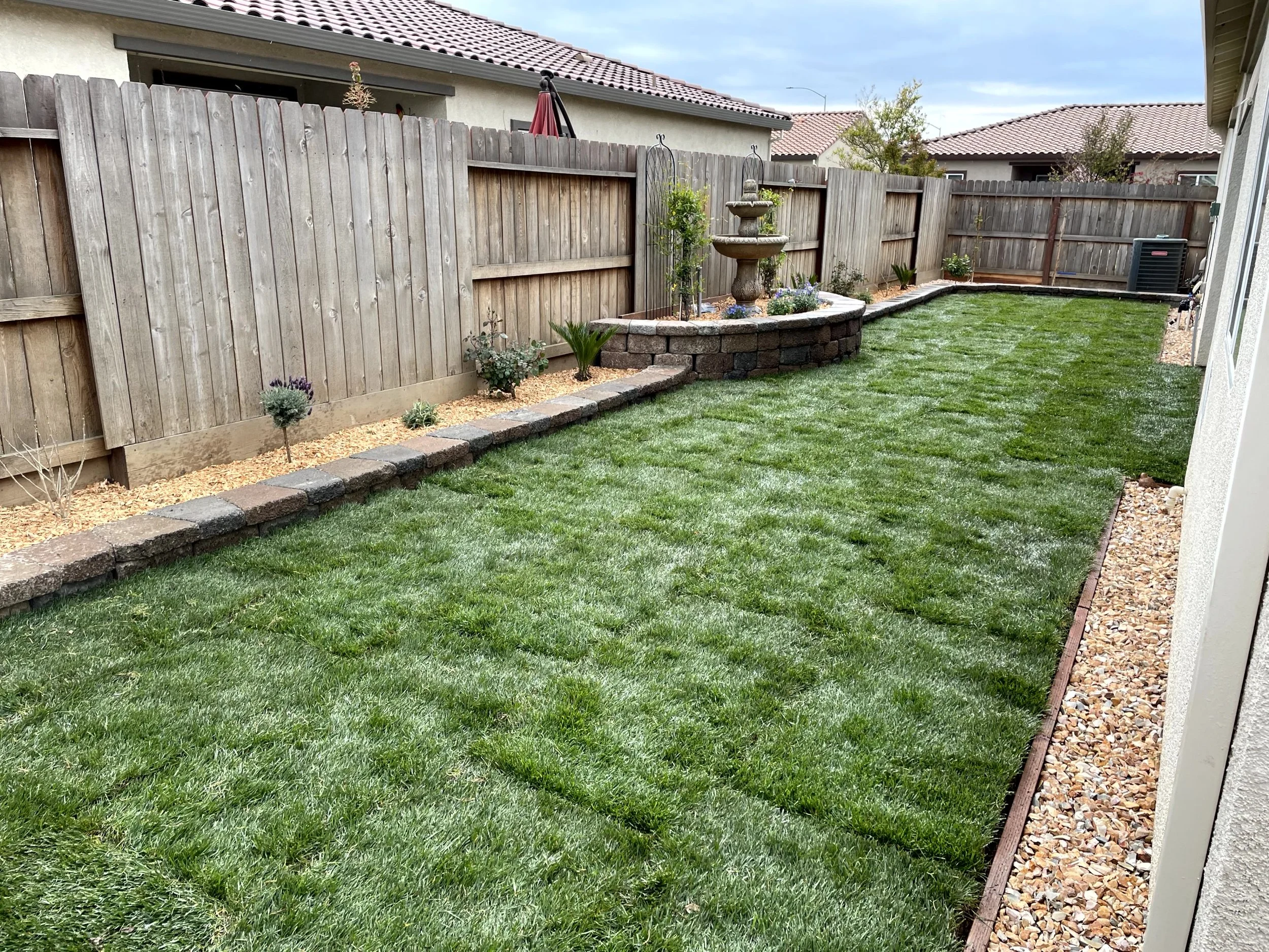 Backyard with a well-maintained green lawn, a wooden fence, a small flower bed with plants and flowers, a stone garden border, and a decorative stone fountain in the corner.