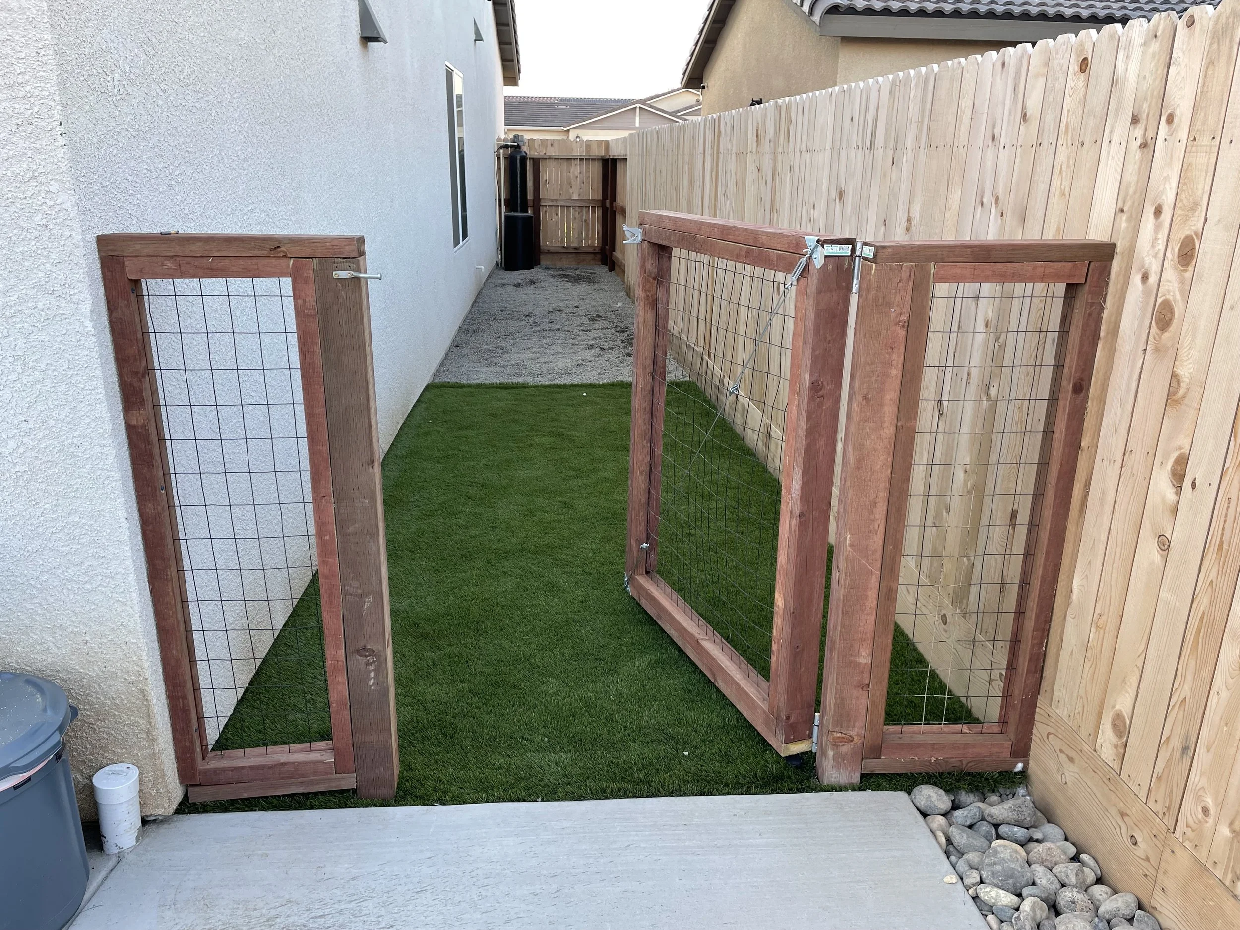 Small backyard with a partial wooden and wire fencing gate, artificial grass, concrete patio, and a gravel pathway, enclosed by house wall and wooden fence.