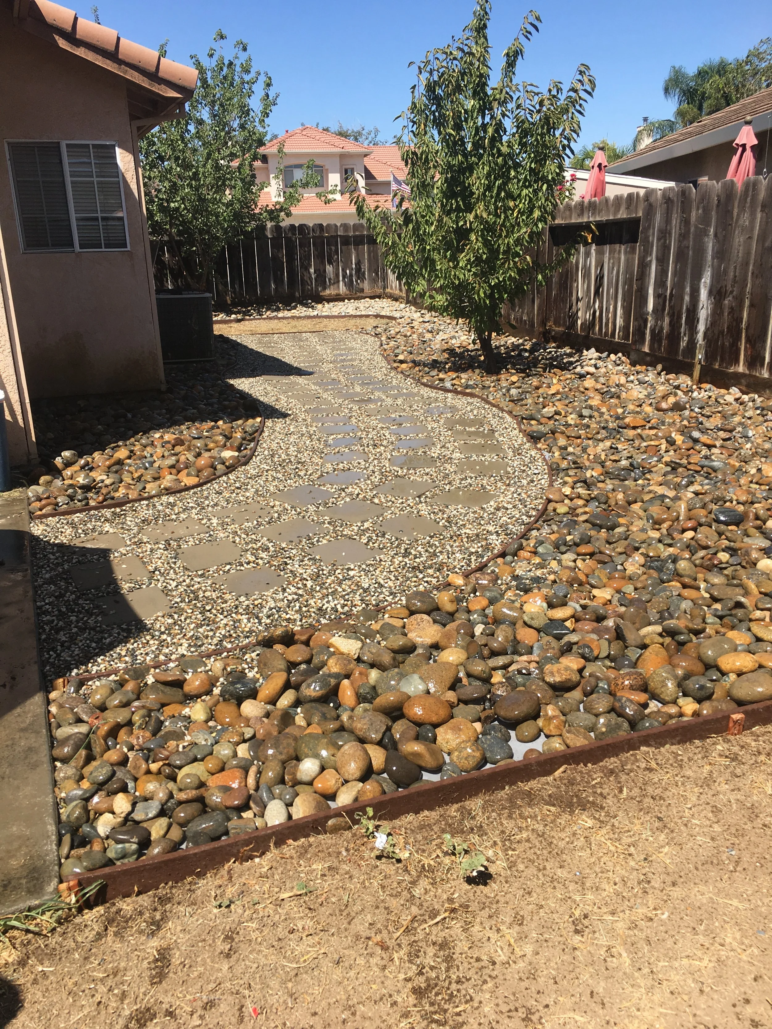 Backyard with a pebble pathway, a small tree, a wooden fence, and patio umbrellas in the background.