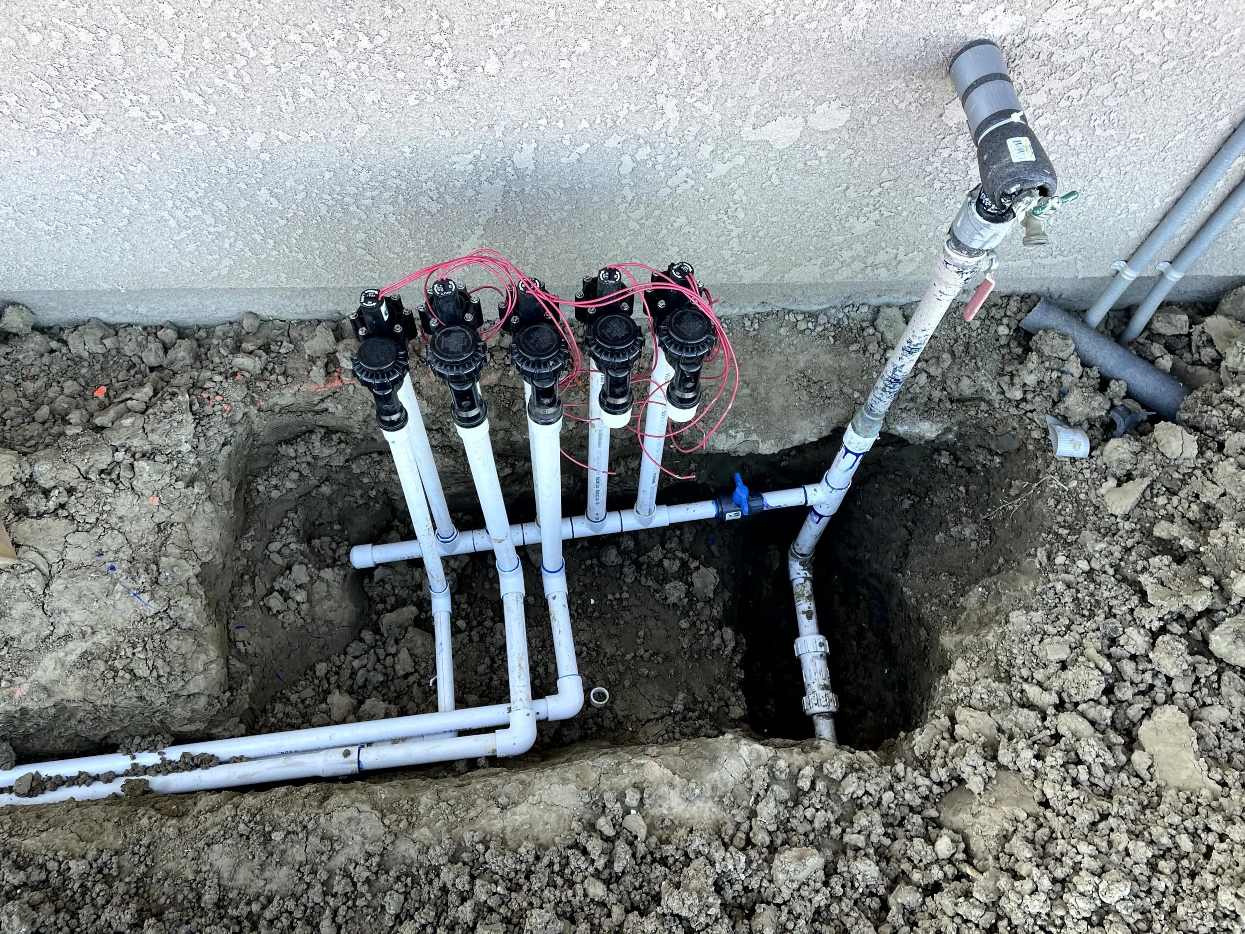 Set of white PVC pipes and fittings connected in a plumbing system in an excavation hole next to a wall, with red wires attached to the valves.