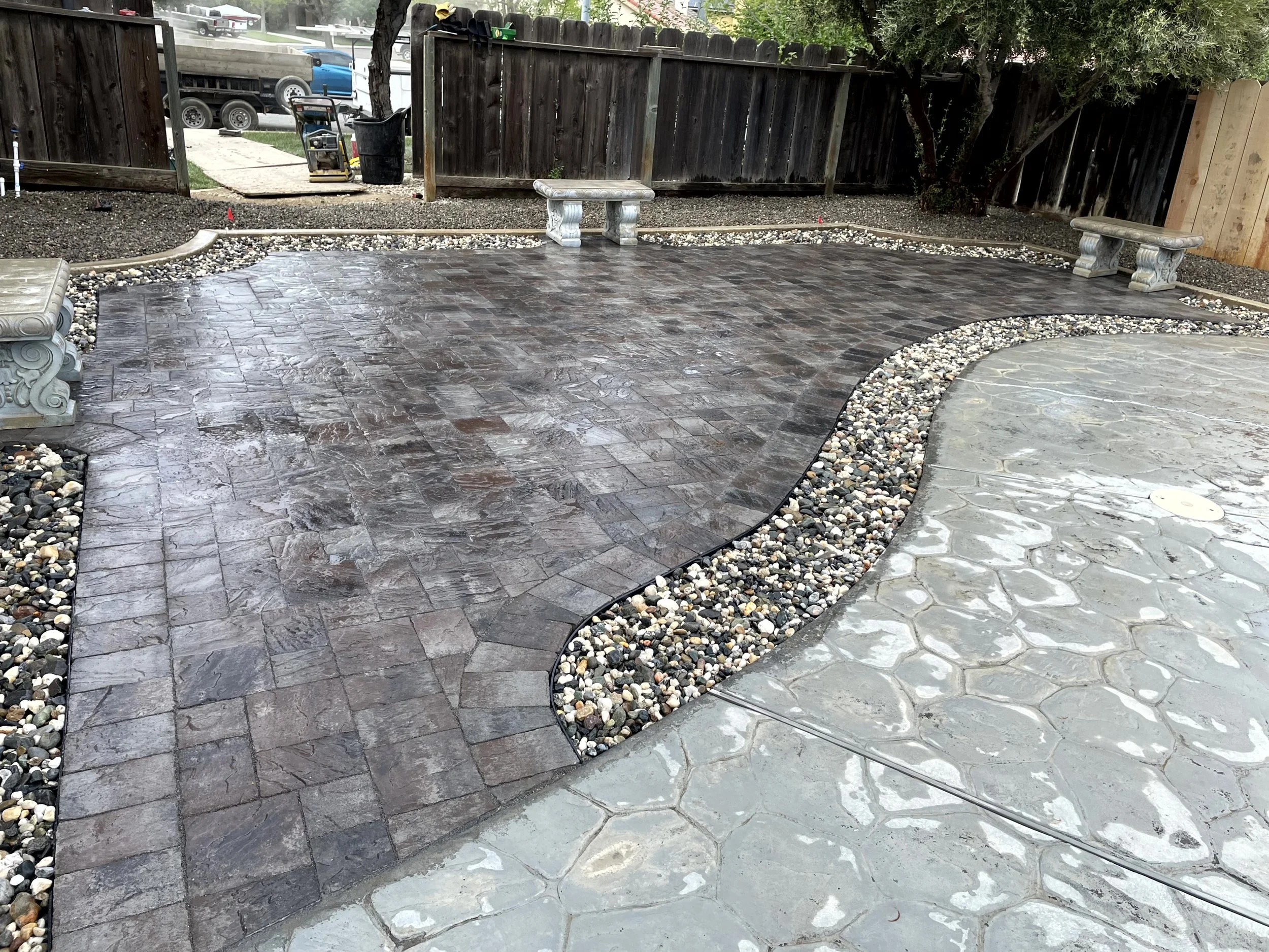 A backyard patio with intricate concrete and brick paving design, including two benches and a surrounding gravel border.