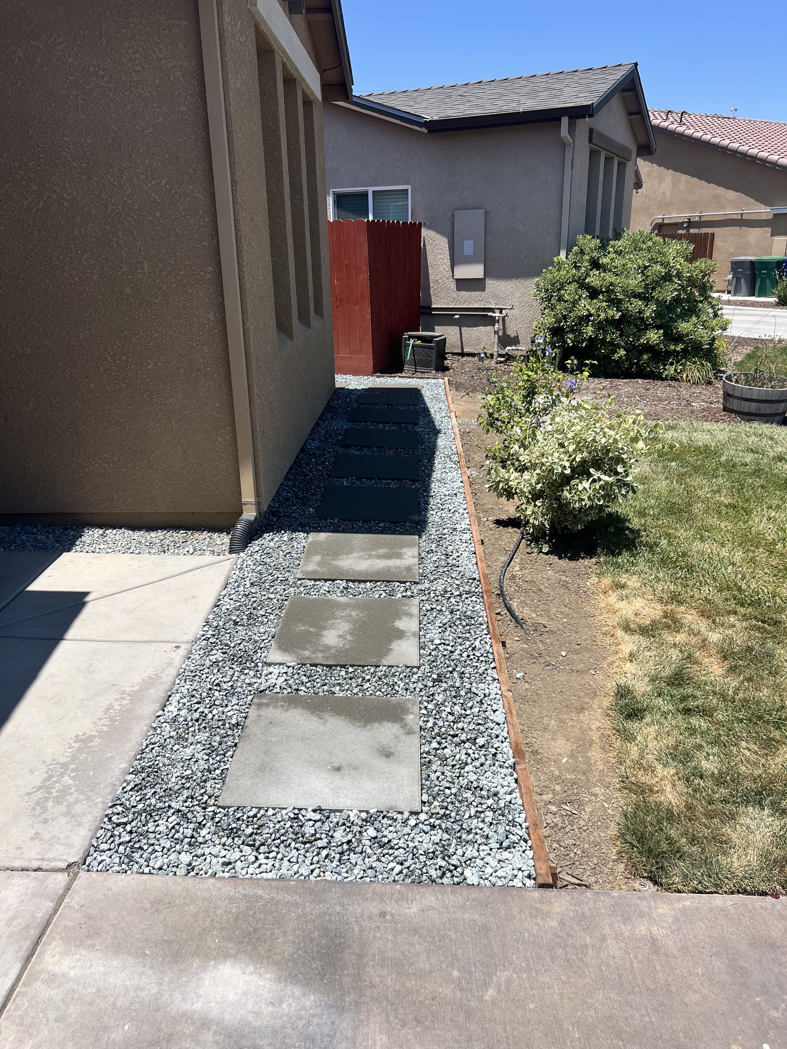 Side yard pathway with stepping stones and gravel, adjacent to a house with beige walls, and a grassy yard with bushes and plants.