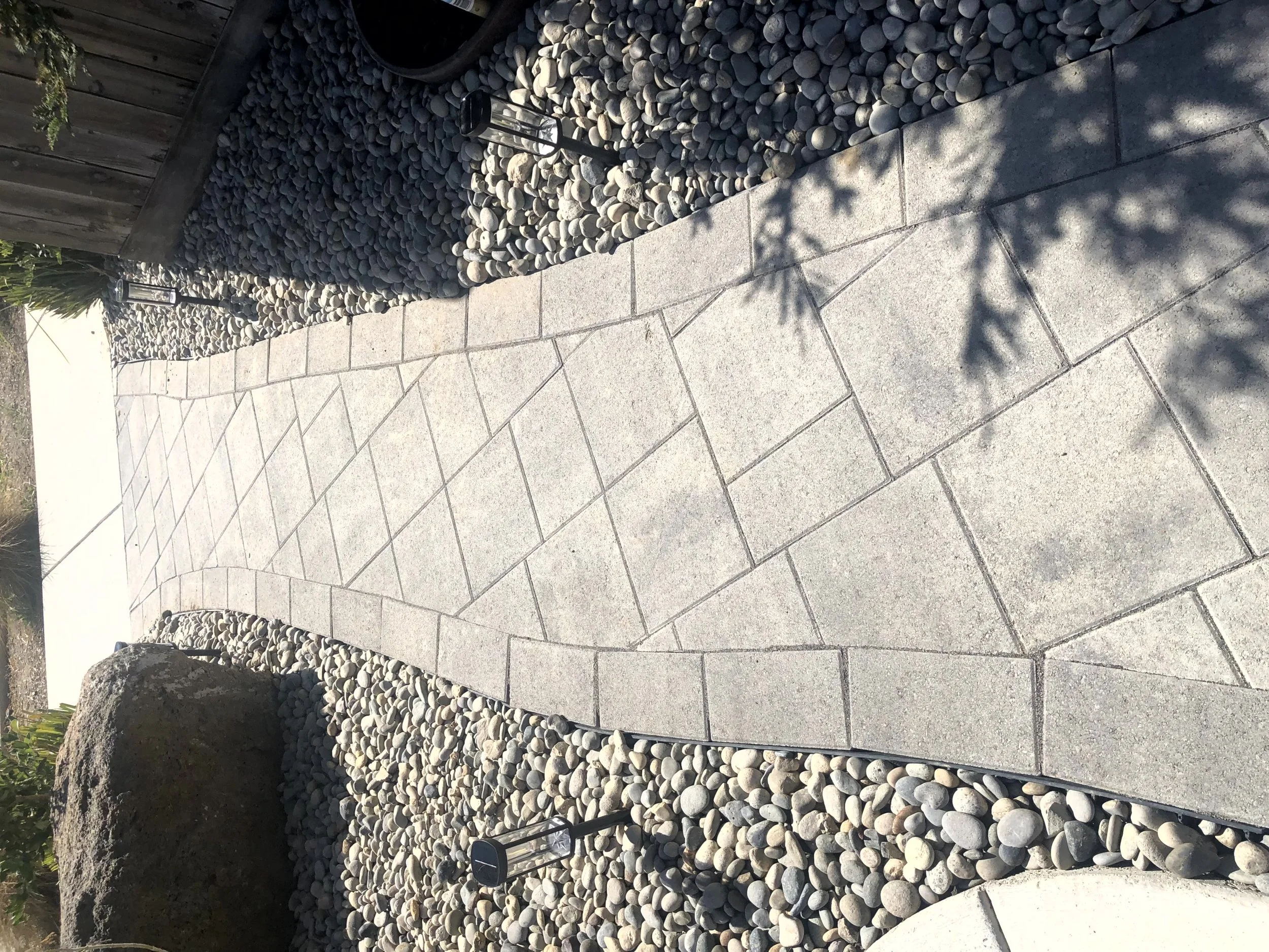 A curved concrete sidewalk bordered by a garden with river rocks and some plants, with a wooden fence and small solar lights along the edge.