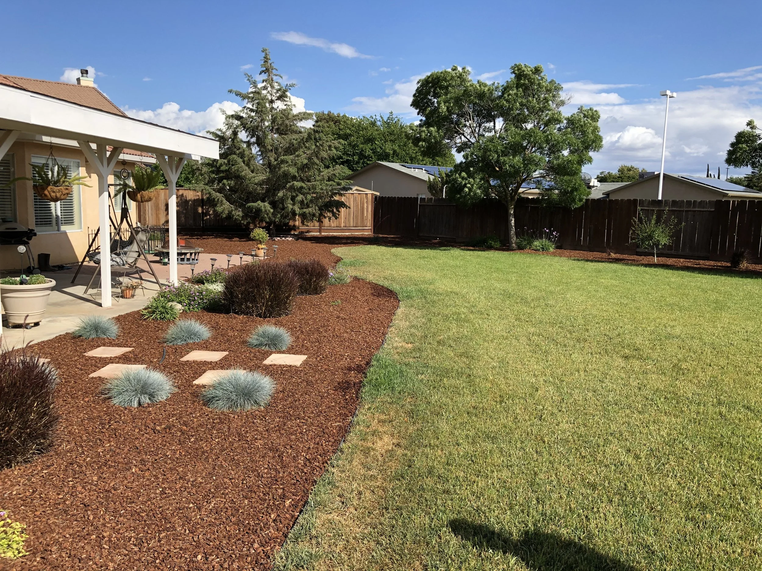 A backyard with a grassy lawn, a shade tree, and a fenced perimeter. There is a covered patio area with hanging plants and outdoor furniture, surrounded by landscaped flower beds with mulch, rocks, and small shrubs.