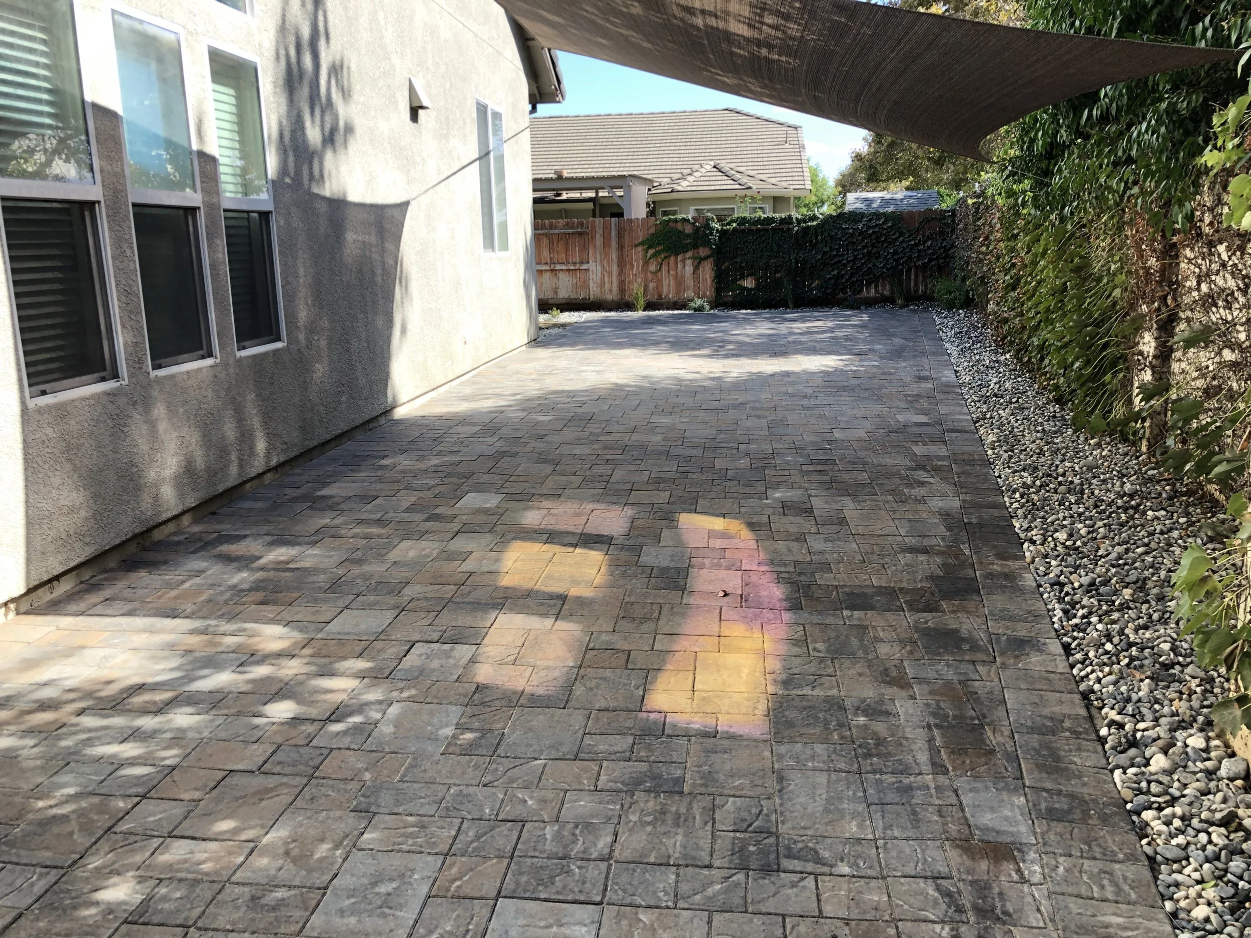 Backyard patio paved with interlocking bricks, bordered by a pebble strip with greenery on the right, and shade from a shade sail overhead. The house wall with windows is on the left, and a wooden fence is at the far end of the yard.