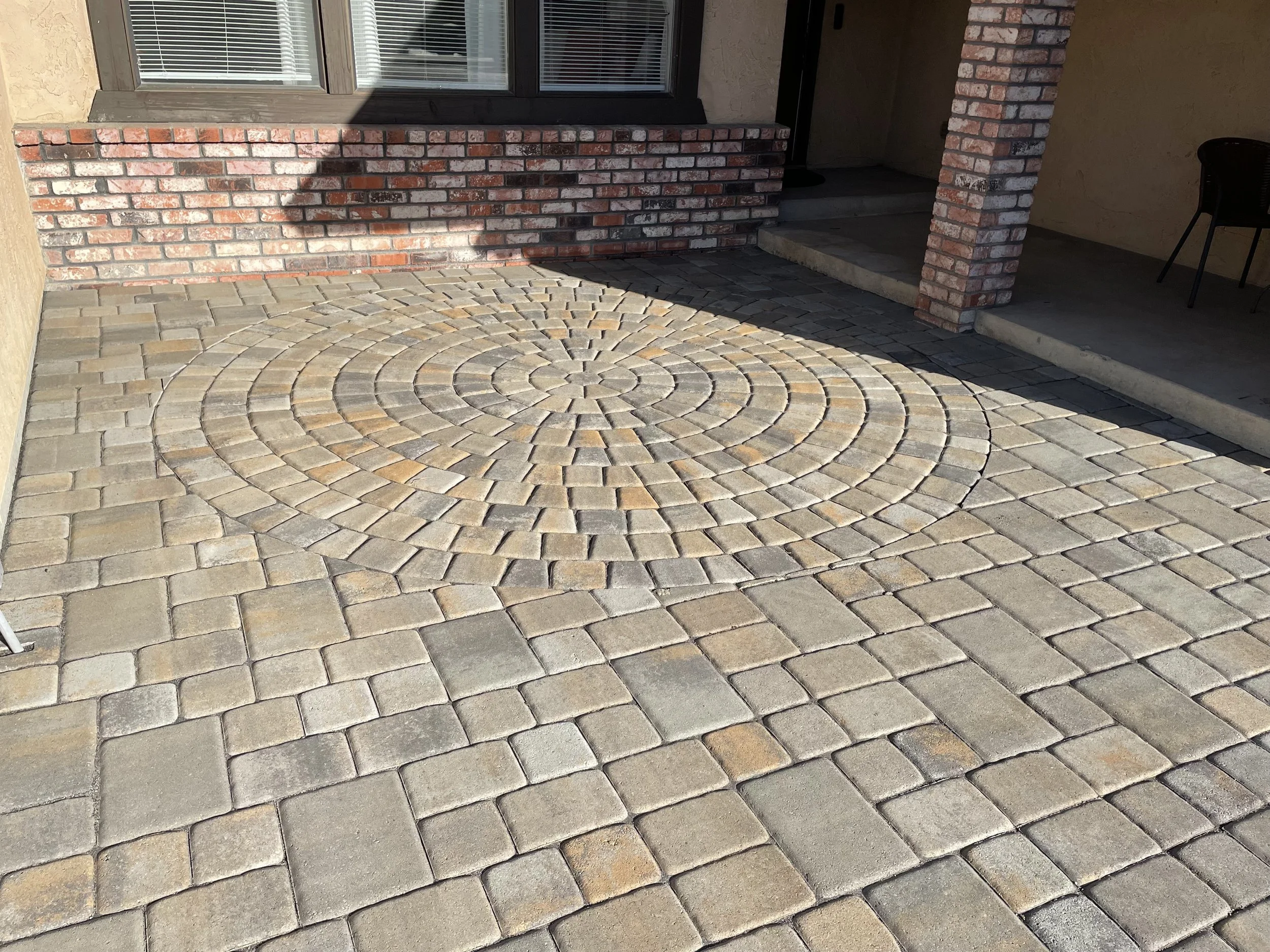 Paved outdoor area with a circular pattern in the center, made of small rectangular bricks, bordered by regular rectangular bricks, next to a brick wall and house entrance.