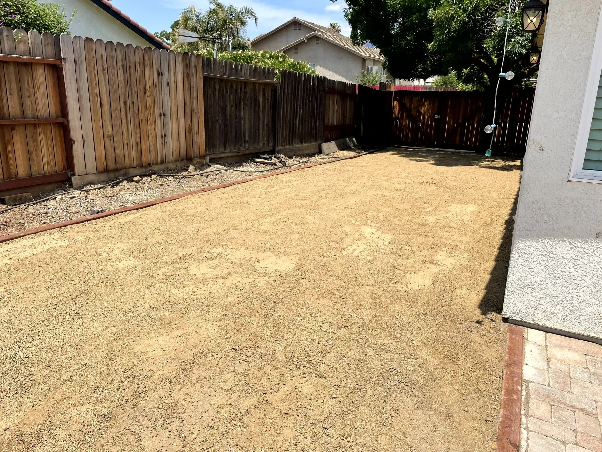 A backyard with a dirt ground, bordered by a wooden fence and a concrete wall on the side of a house, with some trees and houses in the background.