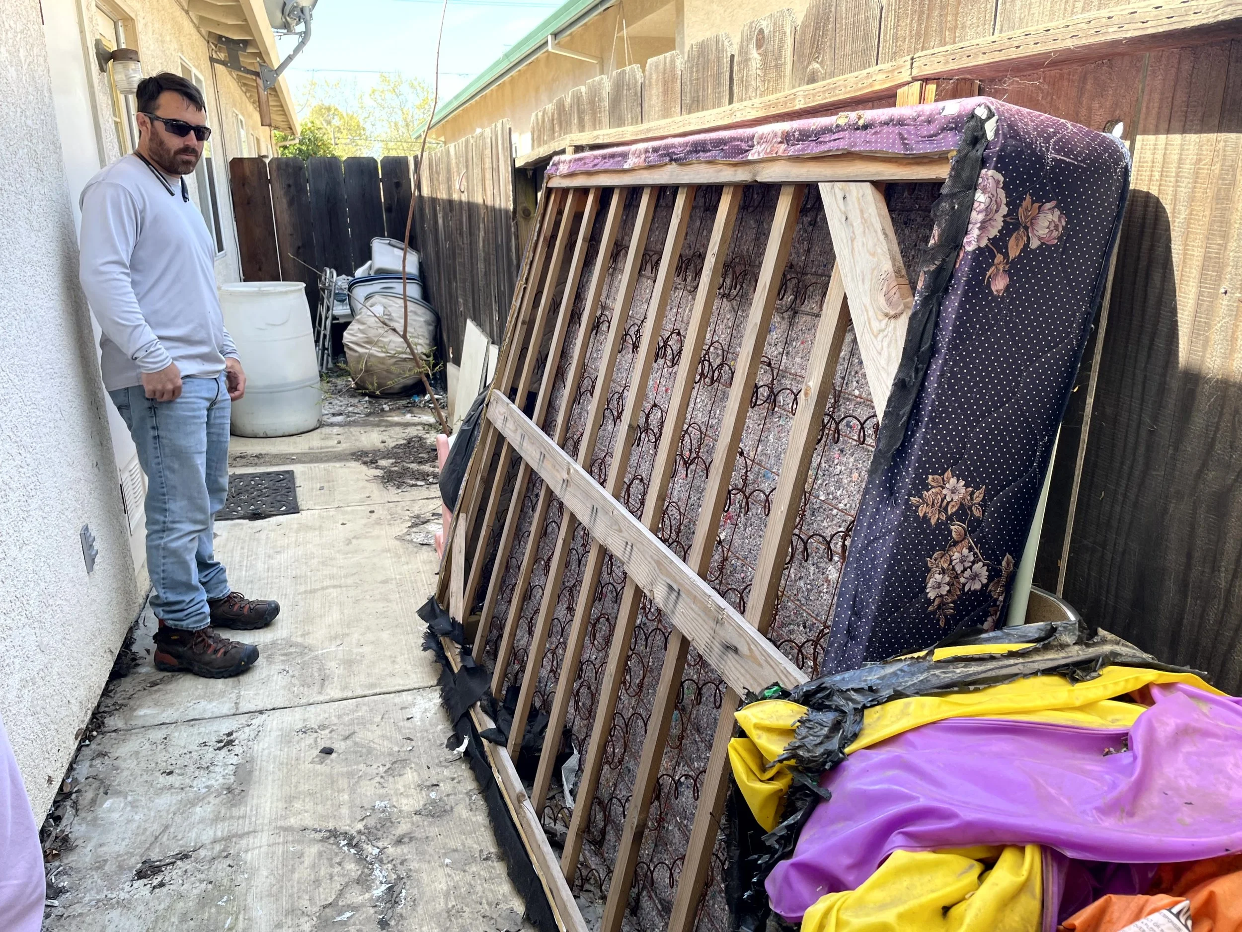 A man with dark hair, beard, sunglasses, wearing a light gray long-sleeve shirt and jeans, standing outdoors next to a brown wooden fence. In front of him are disassembled bed frames, mattresses with floral and patterned fabrics, and various colorful