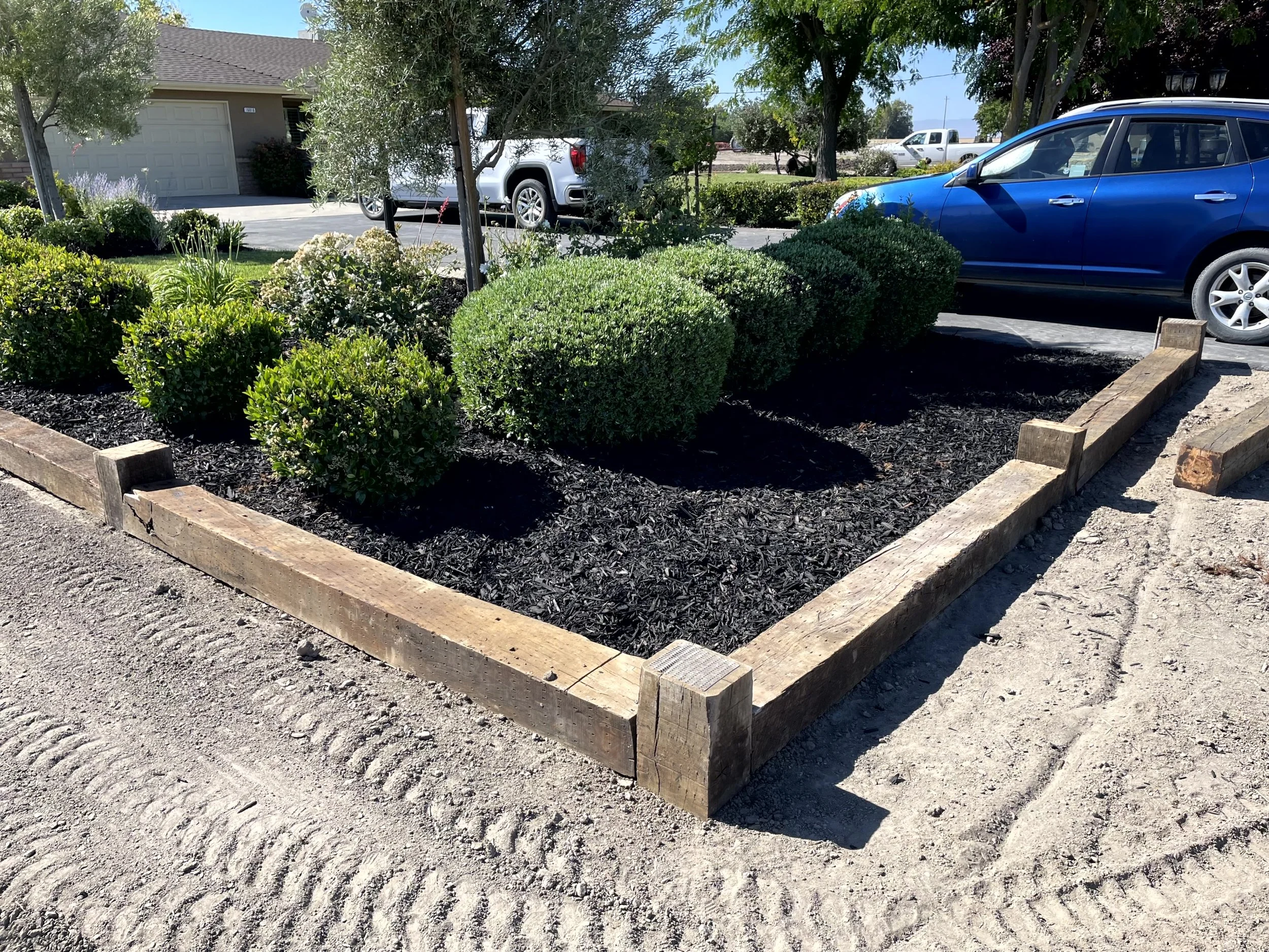 A small landscaped area with green bushes and flowers, bordered by wooden railroad tie timbers, situated next to a parking lot with cars and a house in the background.