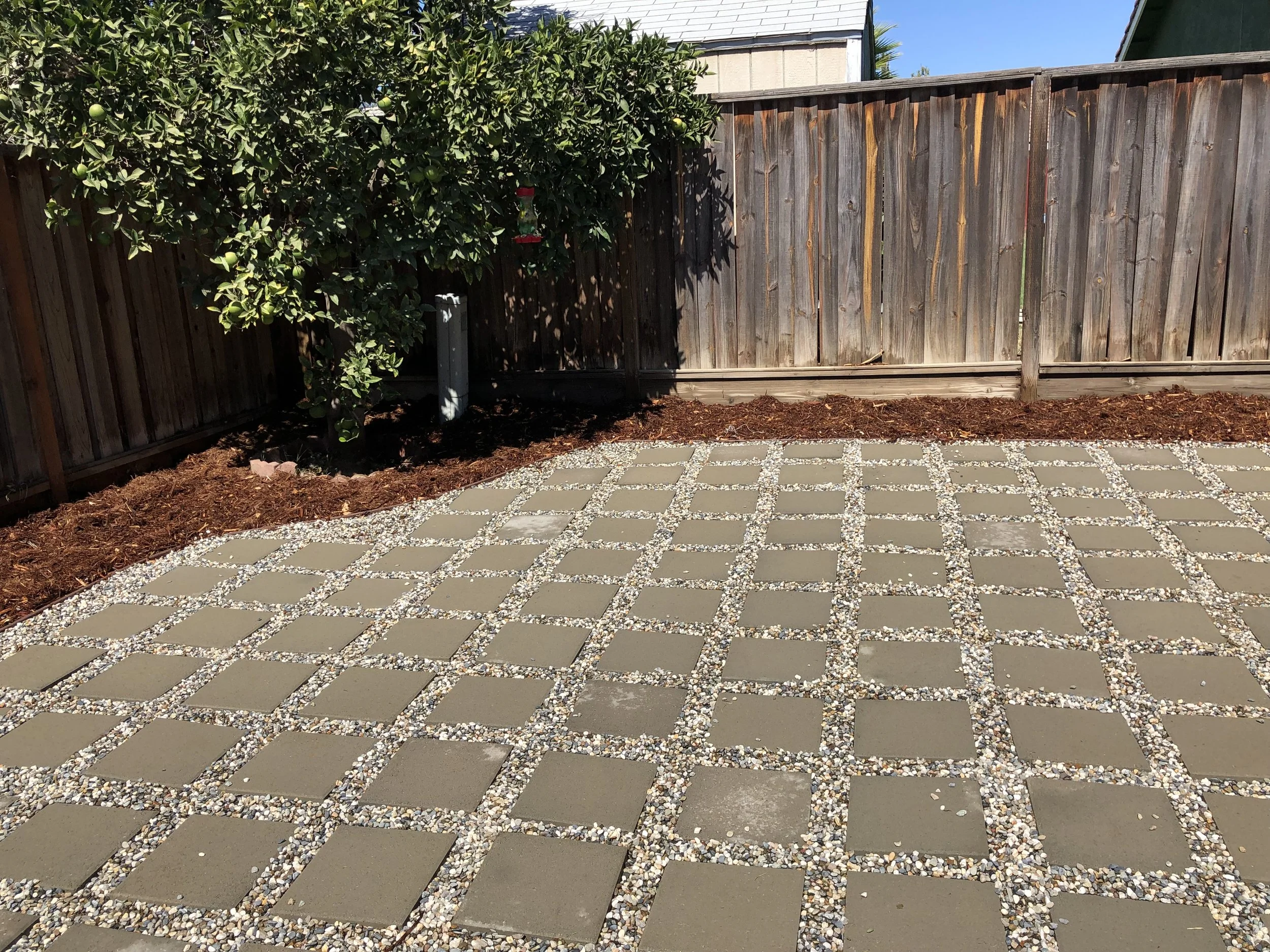 A backyard with a wooden fence, a small tree, mulch, and a patio with concrete pavers separated by small gravel.