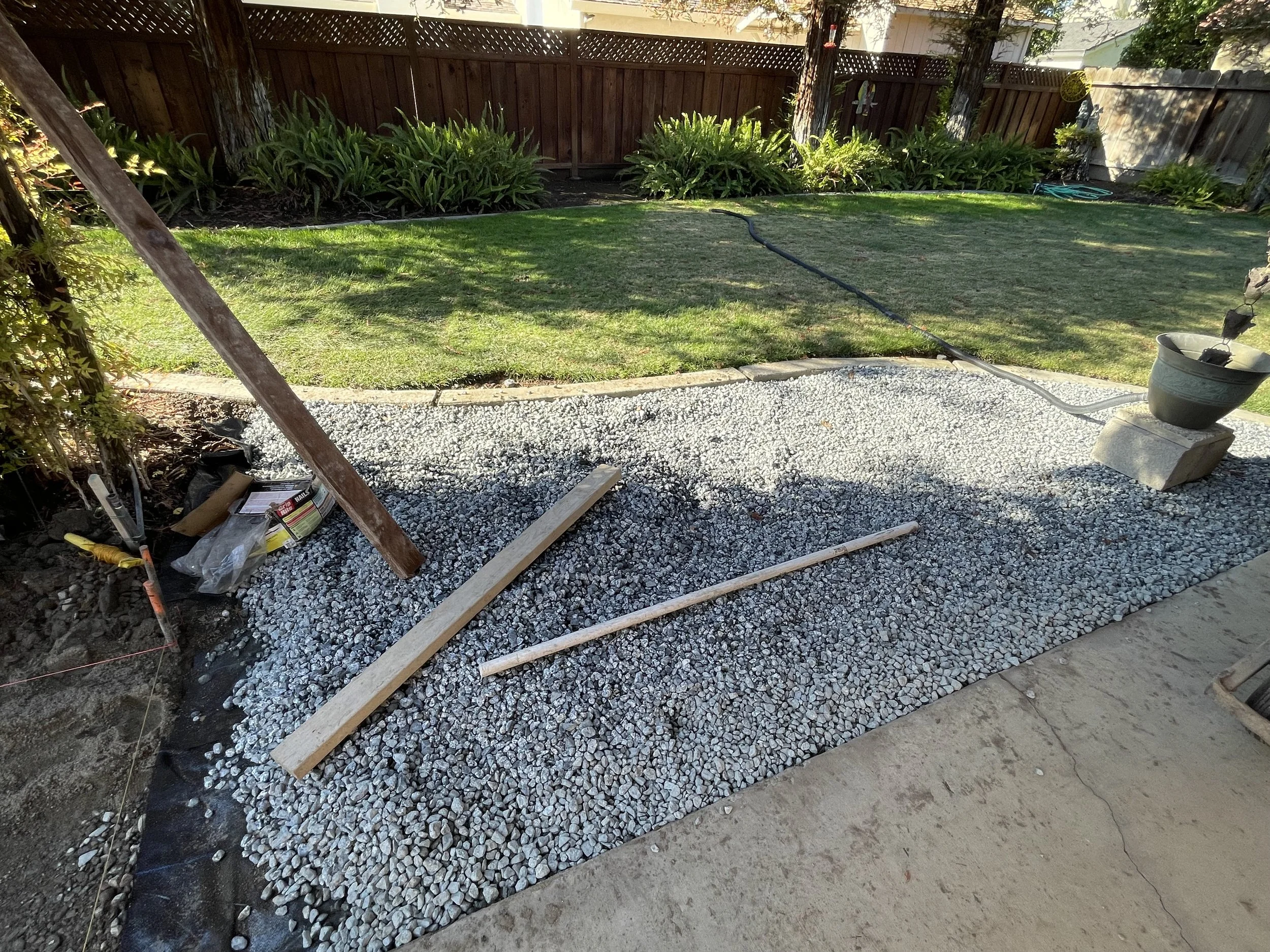 A backyard project in progress showing a gravel pathway being laid, with construction tools and wooden stakes, a grassy area, and a wooden fence in the background.