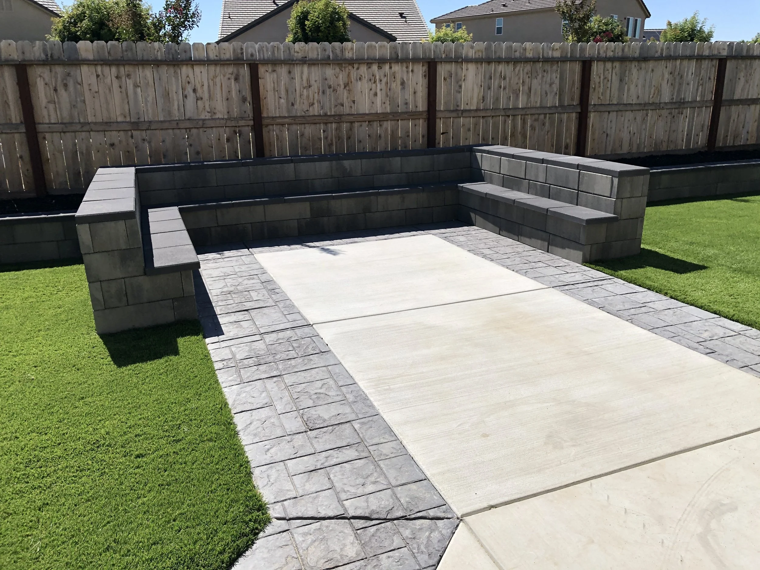Backyard with a patio area made of concrete and stone, with a built-in seating wall and a wooden privacy fence, green grass, and homes in the background.