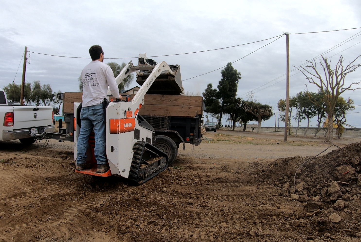 Man operating compact loader on a construction site with a dirt trench and a wooden utility pole in the background.